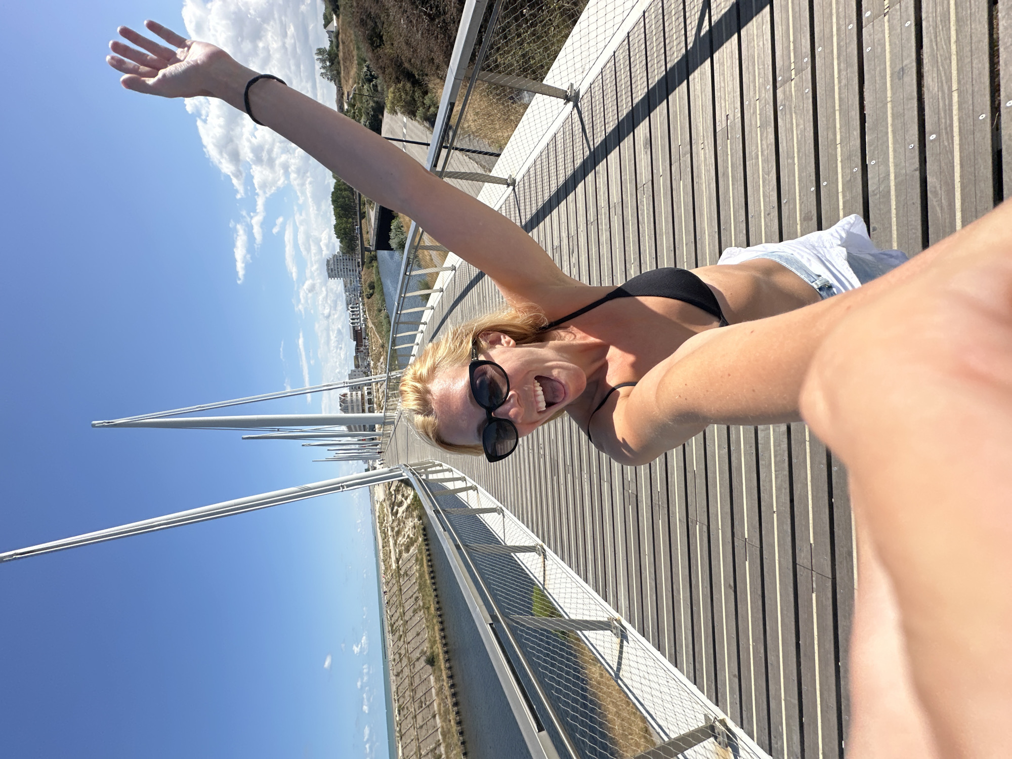 A blonde woman in a black bikini top and white shorts is taking a selfie on a modern pedestrian bridge in Dunkirk, France. She is smiling with her mouth open and her left arm is raised, waving. The bridge has a wooden deck and metal railings, with tall, slender white poles extending upwards. The canal runs alongside the bridge, and beyond that are sandy dunes, beach, and the sea. In the distance, buildings and trees are visible. The sky is blue with scattered white clouds.