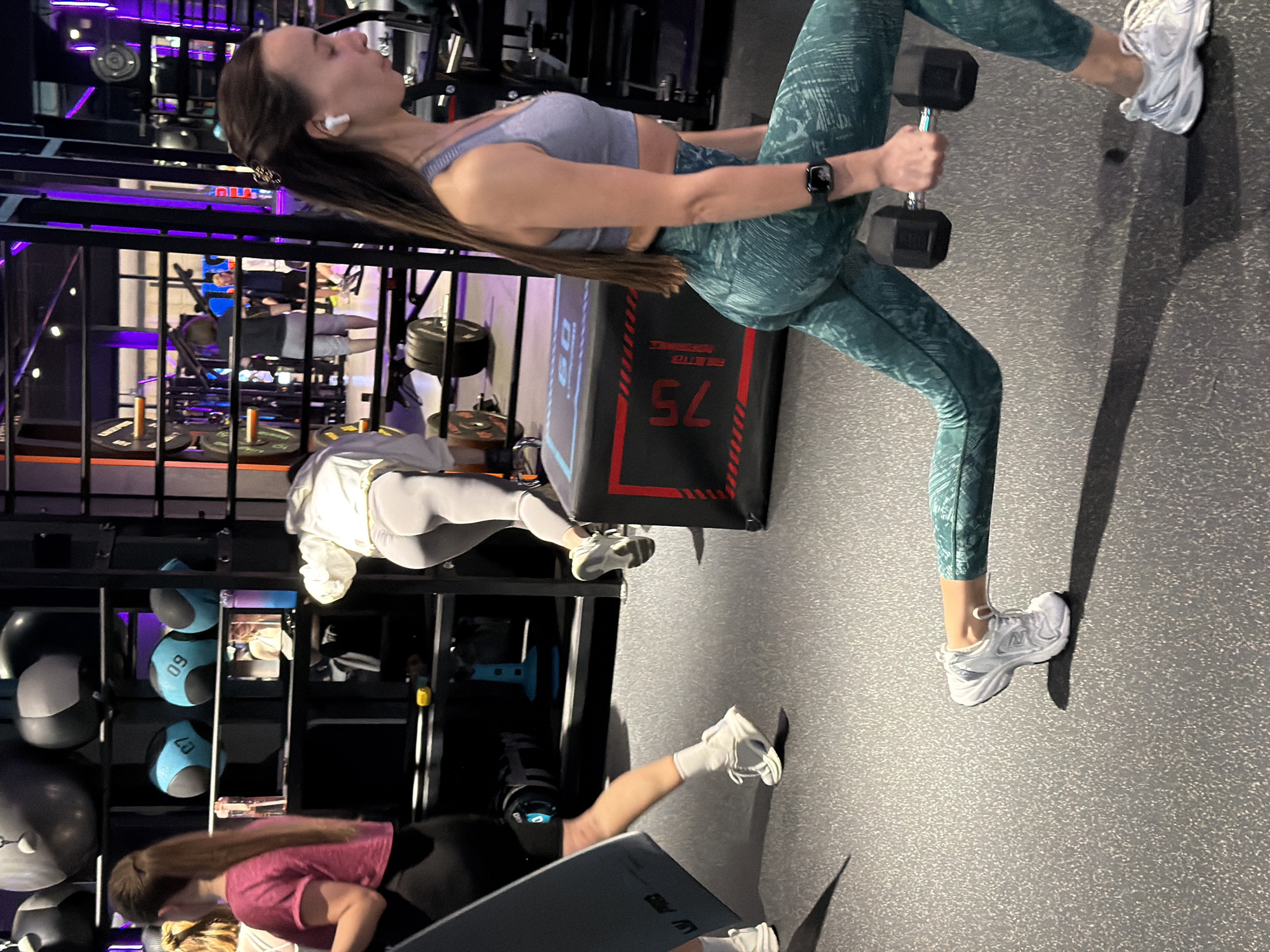 The image shows a gym setting with three individuals. The primary subject is a woman in activewear (sports bra and patterned leggings) lifting dumbbells while positioned on a bench with "75" displayed on it. She wears a smartwatch and wireless earbuds. The second person, in the background, is wearing white athletic clothing and appears to be exercising as well. The third individual is engaged in push-ups on the floor. Gym equipment such as medicine balls and weight plates are also visible. The location is not identifiable as Saint Petersburg, Russia, based on the visual information provided.