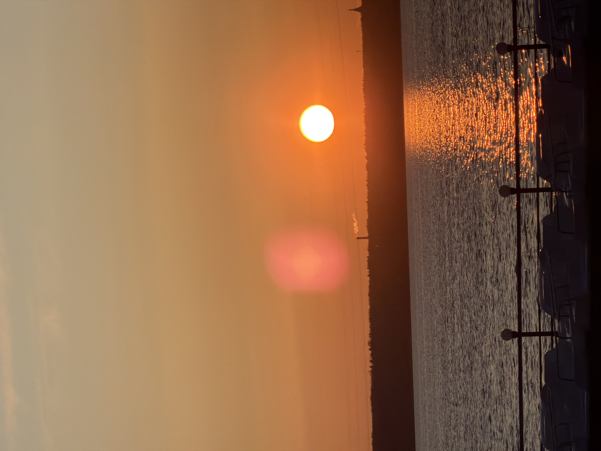 The image captures a serene sunset or sunrise over a large body of water, likely the Izhevsk Pond/Reservoir in Izhevsk, Russia. The sky is illuminated with warm, golden hues, transitioning from a soft, hazy beige to a vibrant orange and yellow. The sun is a brilliant, white-yellow circular disk, prominently positioned in the upper left of the frame (when correctly oriented). A distinct pinkish lens flare is visible below the sun in the sky.
A dark, silhouetted landmass, possibly distant trees or buildings, stretches across the horizon, separating the glowing sky from the expansive water. The water's surface reflects the sunlight, creating a dazzling, shimmering path of bright orange and yellow light that extends from the horizon towards the viewer. The remaining water appears in darker shades of blue-grey, with visible ripples.
In the foreground, on the right side of the image (when correctly oriented), a dark railing or fence structure is visible, suggesting the photo was taken from a promenade, pier, or boat deck. The railing features two distinct posts with spherical tops, resembling lamp posts. The clear sky and warm light indicate fair weather conditions during dusk or dawn. No people or text are visible in this tranquil and picturesque scene.