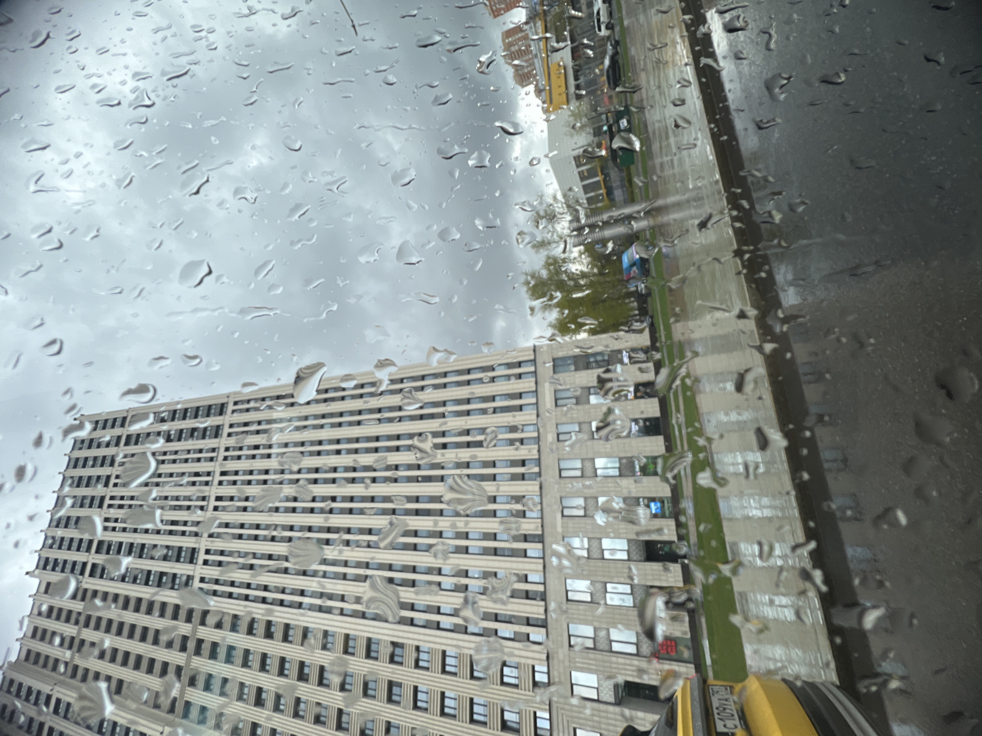 The image, taken through a rain-streaked window, shows a tall building and a street scene. The building is a large, rectangular structure with numerous windows arranged in a grid pattern. The architecture appears modern and functional. The street view includes a sidewalk, greenery, and potentially other buildings or structures further in the distance, but these are blurred and indistinct due to the rain and focus. Part of a yellow taxi is visible at the bottom right corner of the image, and the license plate number is C109TA 197. The general location is the city of Moscow, Russia, based on the vehicle license plate.