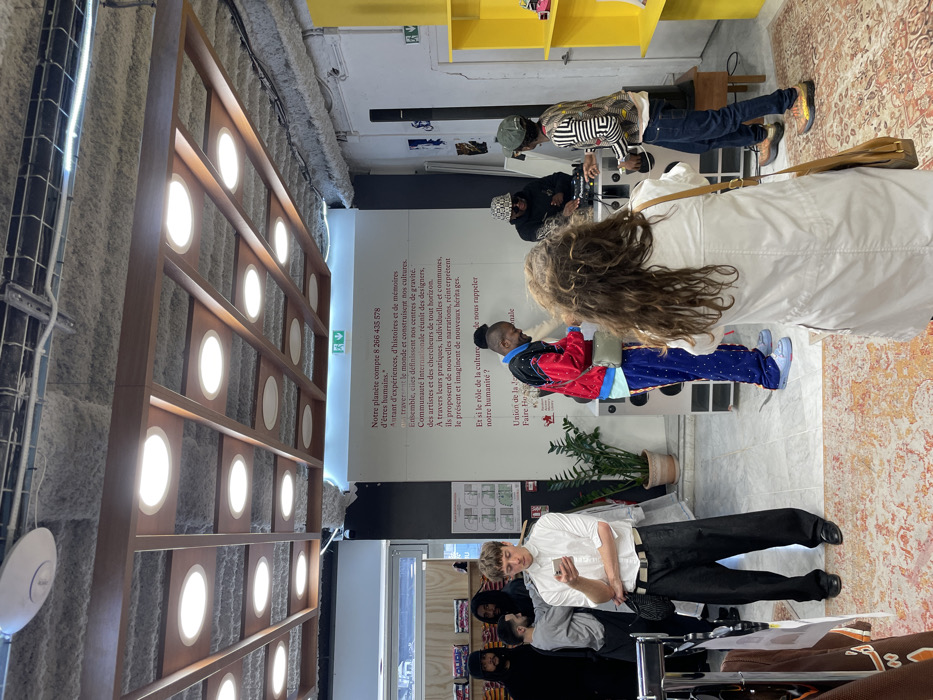 The image depicts an indoor scene within a modern cultural or retail space in Paris, France, as indicated by the French text visible on a large white wall panel. The ceiling is raw concrete, covered with a textured acoustic spray insulation, and features a dark wooden grid structure housing multiple circular recessed lights. Green exit signs are visible on a ceiling beam and a wall.
In the foreground, a woman with long, curly brown hair, wearing a white coat or jacket and carrying a light brown shoulder bag, stands with her back to the camera, holding a device (likely a camera or phone) pointed towards the center of the room. The floor beneath her is covered by an ornate, patterned rug with shades of orange, beige, and red, transitioning to a light-colored floor surface.
The central activity revolves around a DJ setup positioned against the back wall. A male DJ, wearing a patterned bucket hat and sunglasses, is actively operating audio equipment (turntables/mixer) placed on a distinctive silver-colored console with integrated speakers and circular grilles. To the DJ's right, a male wearing a green baseball cap backwards, a geometrically patterned long-sleeved shirt, and dark jeans stands, gesturing with his right hand, seemingly interacting with the DJ. To the DJ's left, another male, dressed in a vibrant red and black patterned jacket, blue patterned trousers, and light blue sneakers, stands looking down towards the console. A potted plant is situated next to the DJ console.
On the far left side of the room, a light-skinned male with short, light brown hair, wearing a white long-sleeved shirt, wide-leg black trousers, and black shoes, stands looking intently at a smartphone held in both hands. A black cross-body bag is slung over his shoulder. Behind him, partially obscured, are several other individuals: two in dark hooded garments and one in a grey sweatshirt. A clothing rack with various garments is partially visible to the immediate left of the man on his phone.
The right side of the room features a bright yellow open shelving unit, partially visible. The overall environment suggests a contemporary event or gathering within a functional, exposed-style interior.