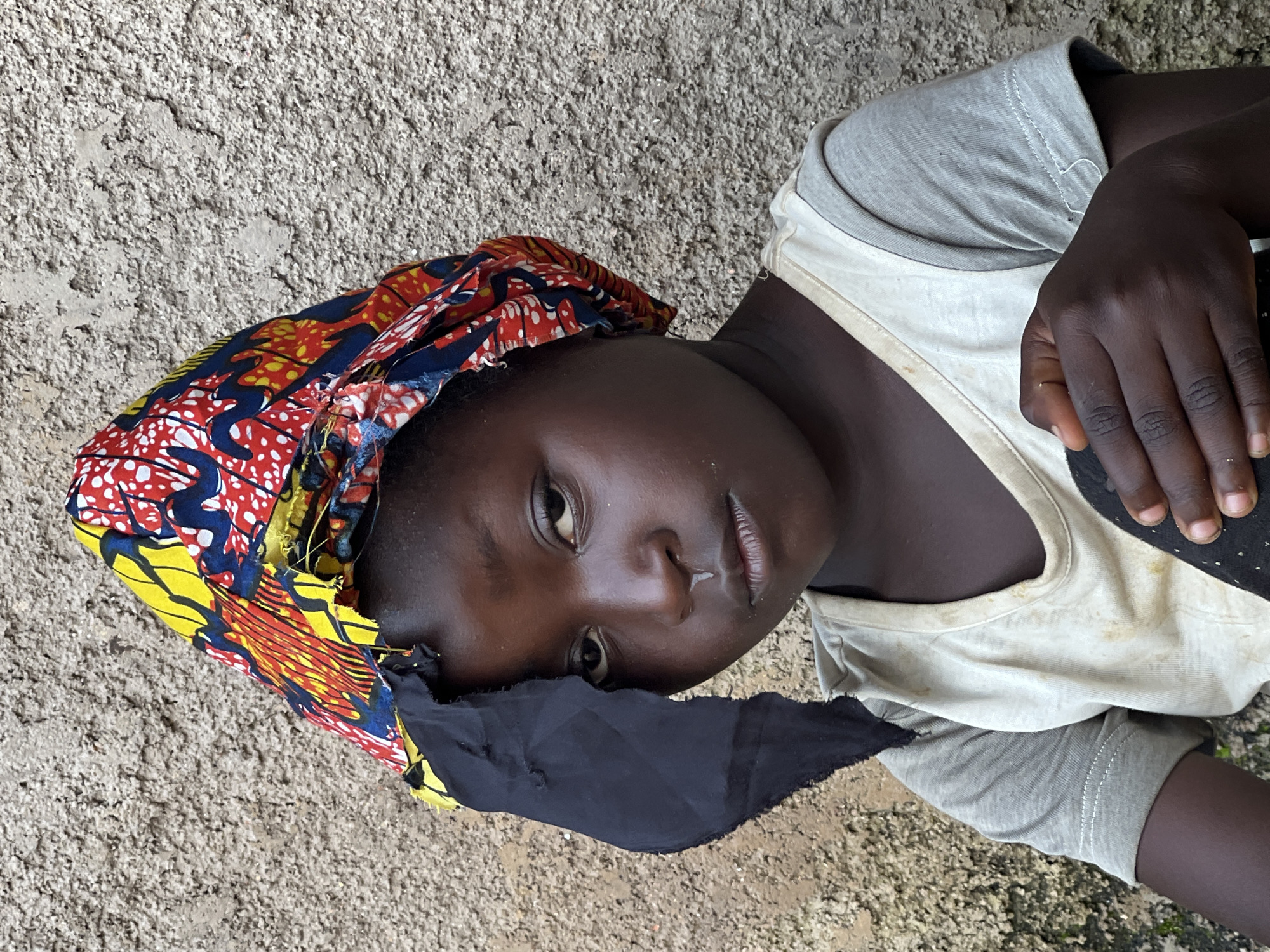 A young girl with dark skin, wearing a gray t-shirt with a lighter t-shirt layered over it, is positioned in front of a textured, light brown concrete wall. Her head is adorned with a colorful headscarf featuring a vibrant print of red, yellow, blue, and white. A piece of black fabric hangs loosely from the side of the headscarf, obscuring part of her face. Her expression is somber, with a slight downward turn of her lips and her gaze directed forward. A noticeable, clear droplet hangs from her nose. Her right hand rests on her chest, holding a piece of black fabric. The lighting suggests an outdoor setting, likely during the day, with the sun casting subtle shadows. The overall scene conveys a sense of quiet contemplation.