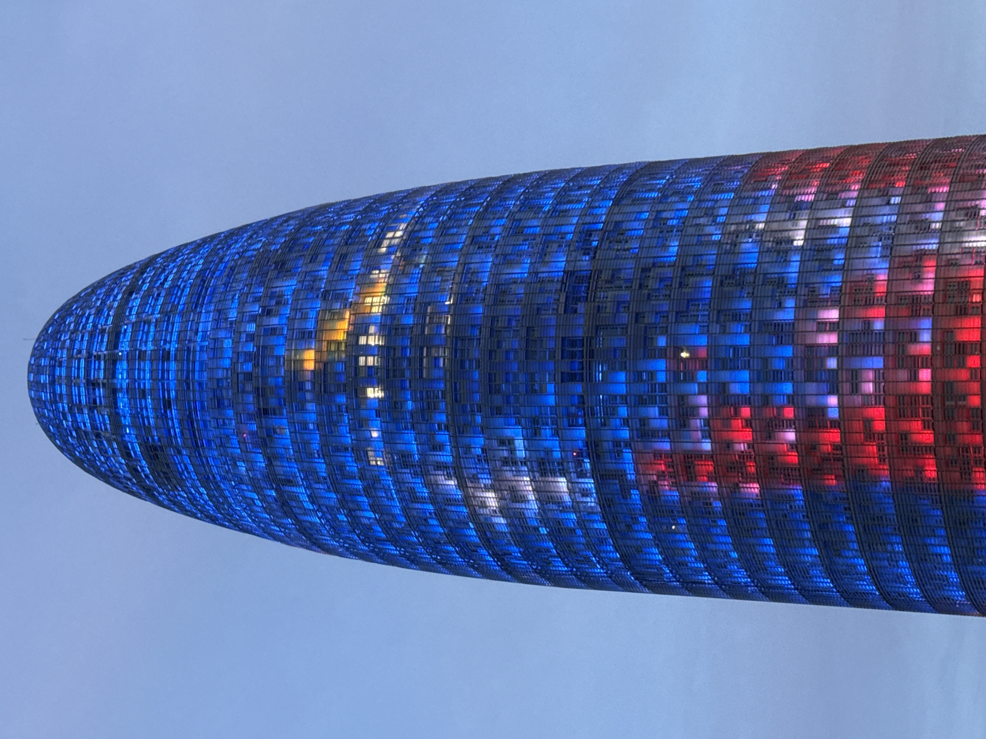 The image displays a vertical, low-angle view of the Torre Glòries (formerly Torre Agbar), a skyscraper located in the Sant Martí district of Barcelona, Spain. The building exhibits a distinctive ovoid, bullet-like, or cylindrical shape, rising prominently against a muted, light blue-gray sky. Its entire facade is composed of a glass and aluminum skin, illuminated by a programmable LED lighting system. The upper and central sections of the tower are predominantly illuminated in various shades of blue, creating a vibrant, patterned mosaic effect across its surface. Towards the lower third of the structure, a substantial portion is lit in a striking red, forming a strong contrast with the blue areas. Scattered throughout the blue and red sections, some individual windows or small groups of windows emit a warmer, yellowish-white light, suggesting interior illumination. No people, vehicles, or other significant objects are visible in the immediate foreground or background, maintaining a clear focus on the illuminated architectural subject. The ambient light of the sky suggests the scene is captured during twilight or early evening.