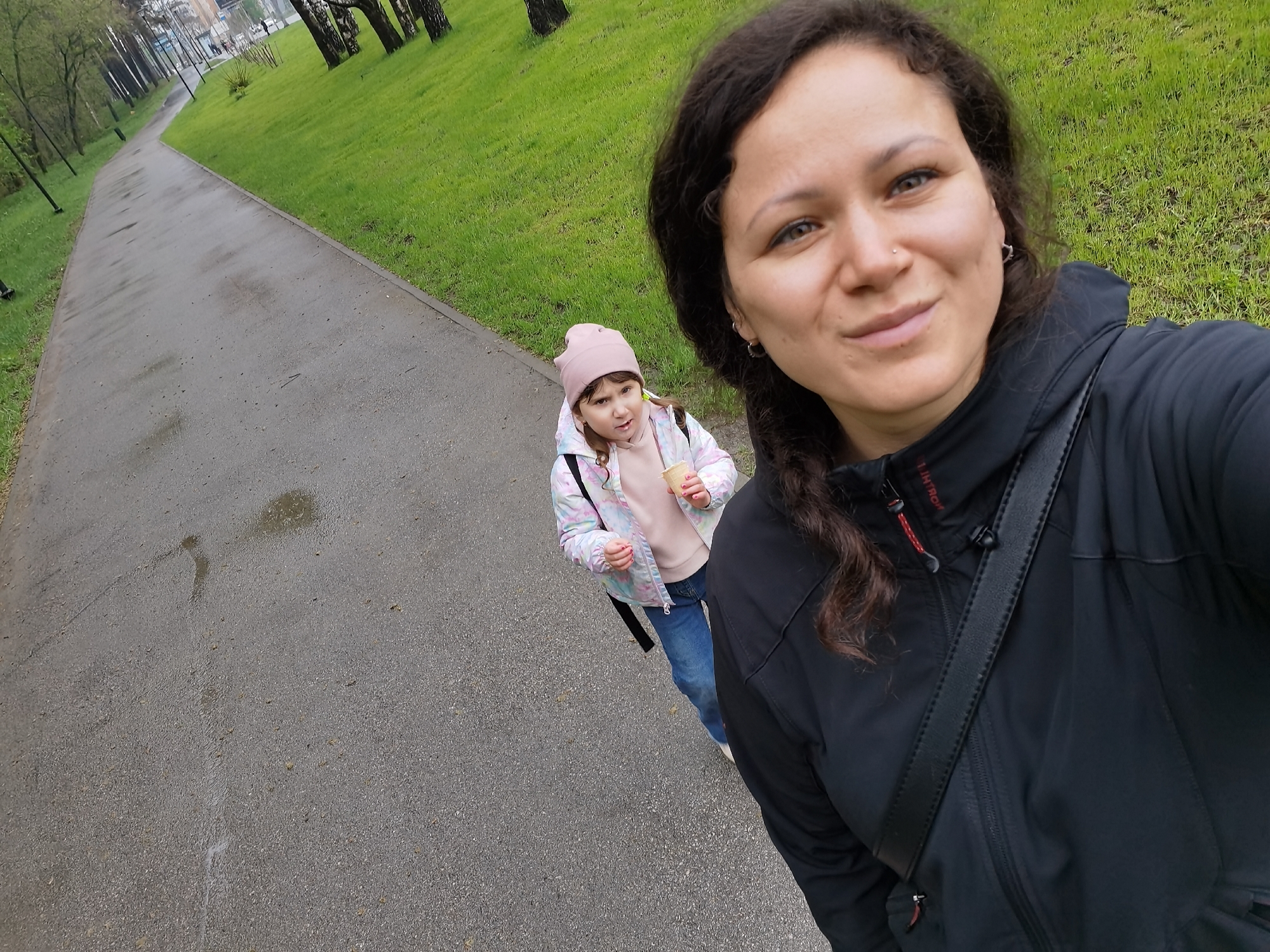 A woman and a young girl are standing on a gray paved pathway in what appears to be a park or green space. The woman, in the foreground, is wearing a black jacket and has dark curly hair. A black bag strap is visible across her chest. The young girl is slightly behind her, wearing a pink hat, a light-colored patterned jacket, and blue jeans, holding a small cup or container. The pathway stretches away into the distance, flanked by green grassy areas with trees. Based on the urban landscape and the presence of birch trees, it is plausible that the location could be within the city of Perm, Russia.