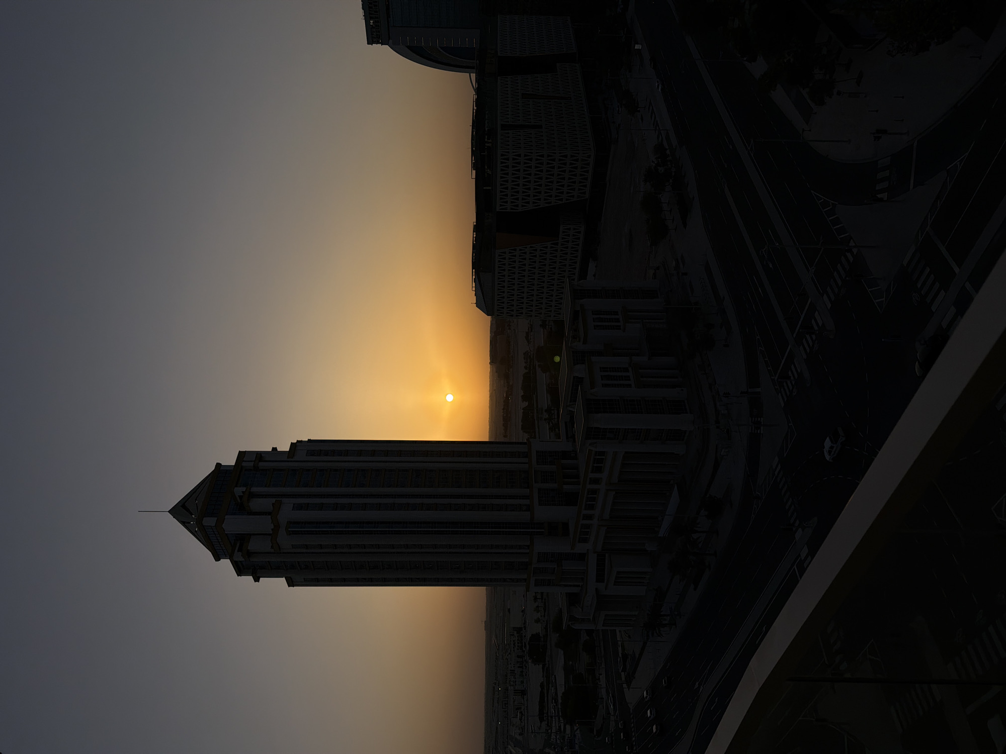 A silhouette of a tall skyscraper and two modern buildings are visible against a gradient sunset sky in an urban environment. The sun, a bright orange orb, is positioned slightly to the left of the center of the image, casting a warm glow. The skyscraper, with its distinctive tiered design and pointed top, dominates the left side of the frame. To its right, two lower, block-like buildings with intricate geometric patterns on their facades are visible. In the foreground, a dark, angled structure, possibly a balcony railing, partially obscures the view. Below the buildings, a network of roads with streetlights and crosswalks is discernible, with a few cars sparsely distributed. The overall scene suggests dusk in a modern city, with the architecture and infrastructure hinting at a developed metropolitan area.