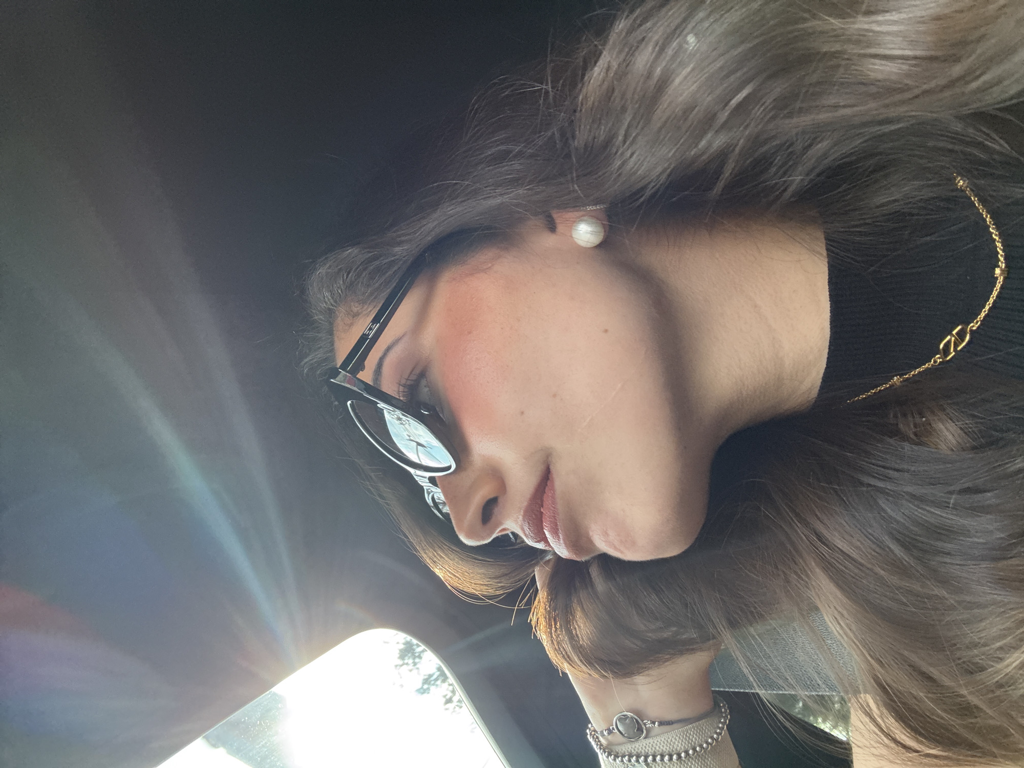 A woman is positioned within a vehicle, likely in the driver's seat, as indicated by the presence of sunlight shining through what appears to be the windshield on the left side of the frame. She is wearing sunglasses and a pearl earring. A gold necklace is visible, as well as bracelets on her wrist. The scene is enclosed within the car's interior, suggesting the location is a parked or moving vehicle within an urban environment. The specific location in San Bernardino Tlaxcalancingo, Mexico cannot be determined from the image alone.