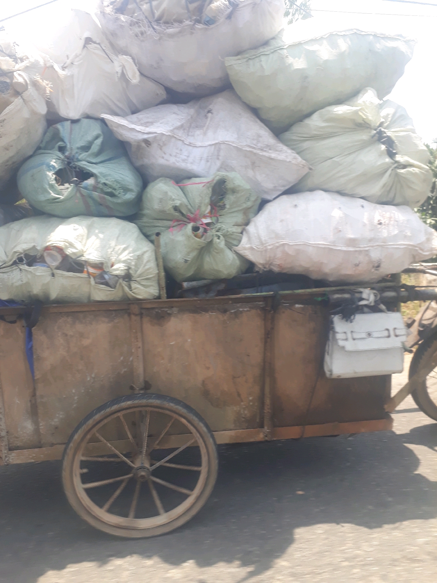 A close-up, eye-level shot captures the side of a cart overflowing with large, tightly packed sacks, suggesting a scene of transport or collection of goods, possibly recyclables, on a street in Pangkalpinang, Indonesia. The cart itself appears to be made of weathered metal, with a prominent, spoked wheel on the left side. The sacks are varied in color and size, with some being a light green and others a pale white or grey, and they are secured with what appear to be ropes or ties. The sheer volume of the sacks implies a significant amount of material. The lighting suggests it is daytime, with bright light casting shadows and creating highlights on the sacks. The background is blurred, with hints of foliage and a bright sky, but the focus remains on the cart and its contents, indicating a snapshot of a common logistical process in the city.