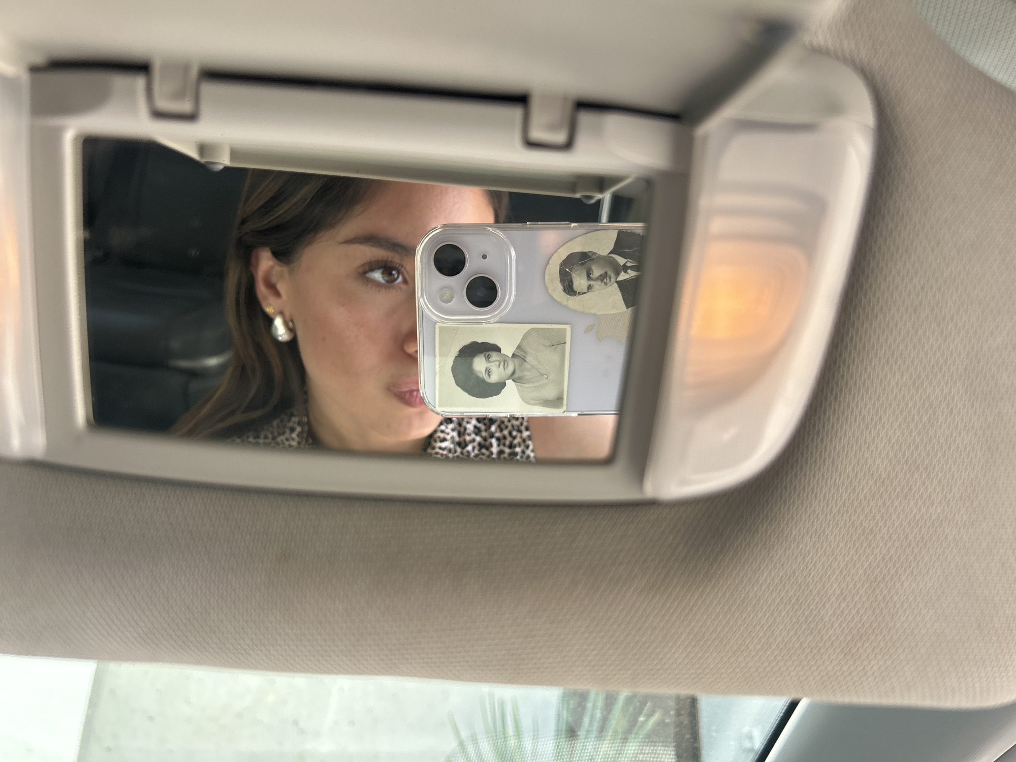 A woman is seen in the reflection of a car's vanity mirror. She is holding a phone in front of the mirror, capturing her reflection. The phone has two vintage photos attached to its case. The car's visor, the mirror itself, and a portion of the car's interior are also visible. The location in the city is Unknown, Unknown.