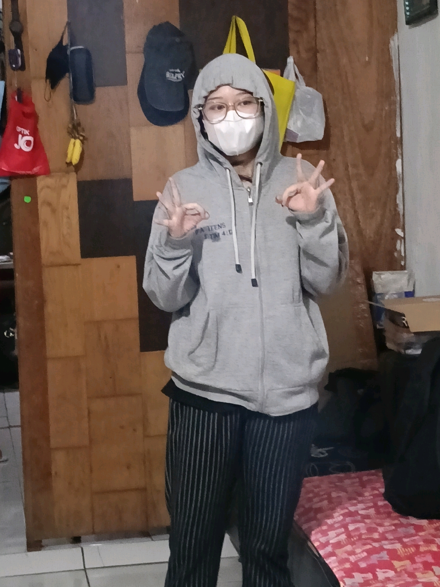 The image shows a full-body shot of a person wearing a gray hooded sweatshirt, a white face mask, and black and white striped pants. The person is standing in what appears to be a room with wooden paneling on the walls and a tiled floor. On the wall behind the person, there are several items hanging, including a red bag with the word "OPTIK" on it, a dark-colored cap, and a yellow bag. To the right of the person, there is a wooden door and a glimpse of a red and white patterned surface, possibly a bed or blanket, with some boxes. The person is making a peace sign with both hands. The lighting suggests an indoor environment, possibly in a residential setting within Jatimulya, Indonesia.