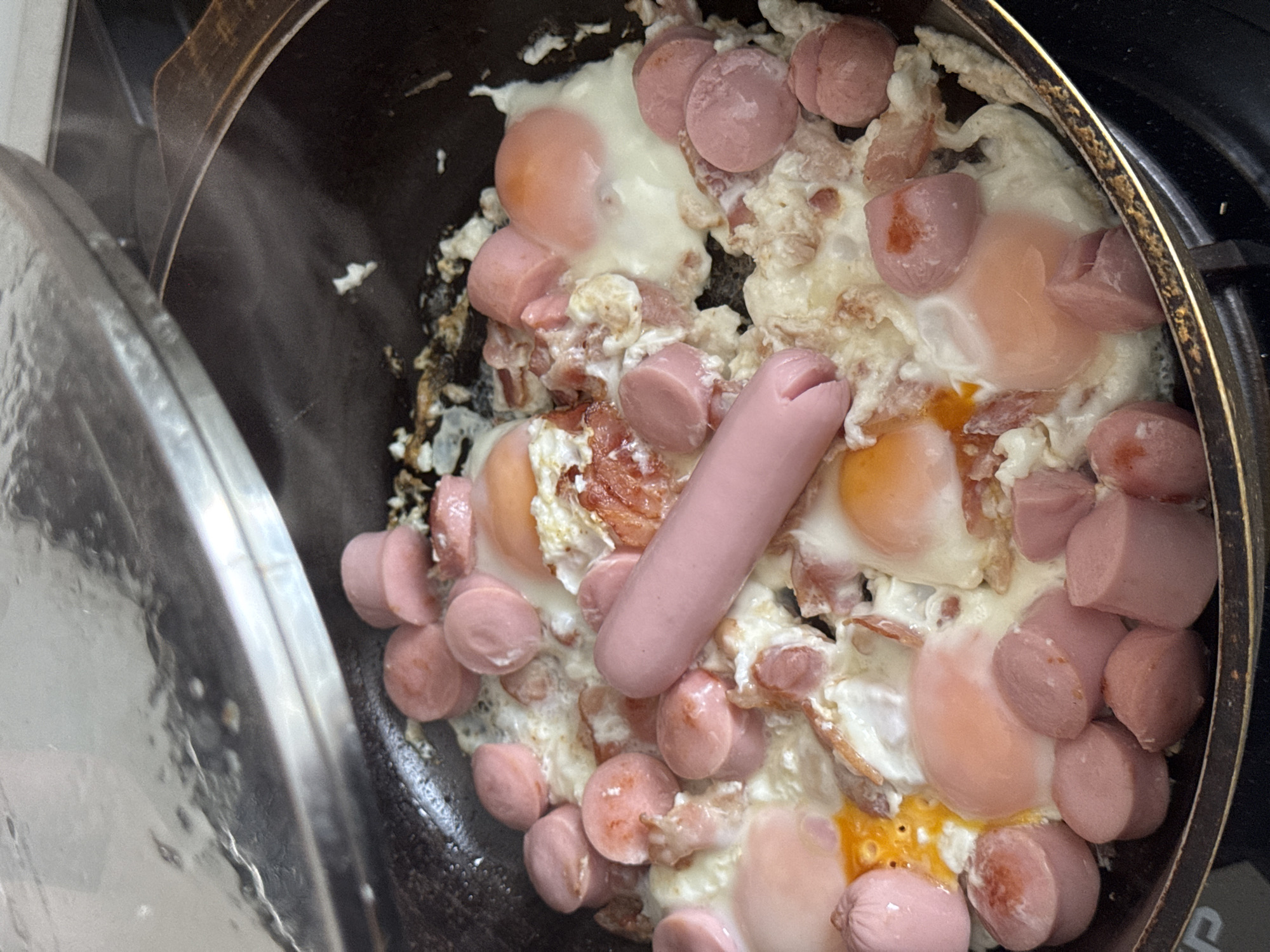 The image shows a close-up, high-angle view of a frying pan filled with food, possibly a breakfast dish. The pan contains several fried eggs with partially cooked yolks, interspersed with many sliced sausages and some pieces of what appears to be bacon. A single, whole, uncooked sausage lies on top of the cooked food. Steam is visible rising from the pan, suggesting the food is hot and actively cooking. The pan itself is dark-colored, likely cast iron or a non-stick material, and is placed on a black stovetop. A reflective metal lid is partially covering the pan, angled upwards and to the left, showing condensation on its underside. The overall scene is domestic and appears to be in a kitchen. There is no discernible information in the image that would allow for the identification of a specific location in the city of Perm, Russia.