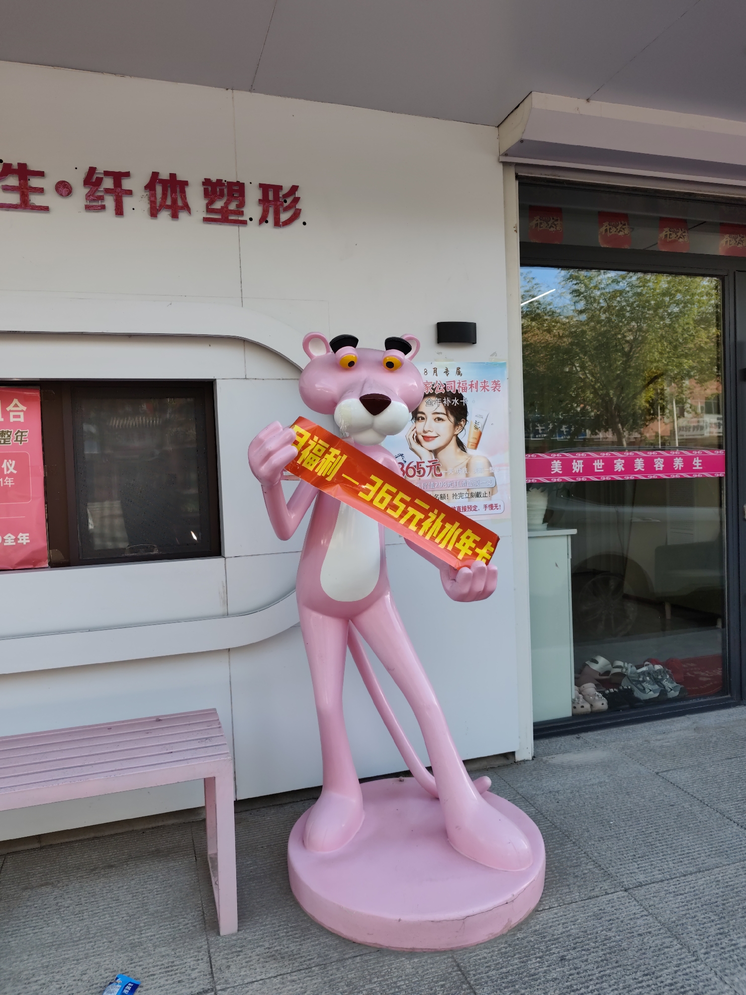 A bright pink statue of the Pink Panther stands outside a business on a paved walkway in Lindian, China. The statue is holding a red banner with Chinese characters in yellow. The business has a white facade with red lettering above a glass storefront. Red decorative elements are visible in the storefront window. A pink bench is situated to the left of the statue, and a poster featuring a woman is partially visible to the left of the statue. Trees and a vehicle are reflected in the glass of the storefront. The ground is a grey, tiled surface.