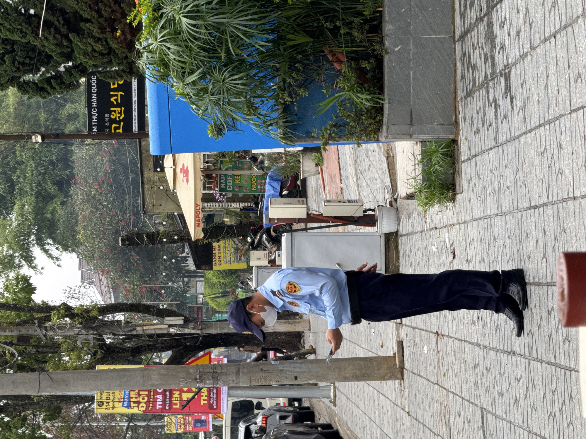 The image captures a street scene in Lâm Viên - Đà Lạt, Vietnam, showing a man in uniform on a sidewalk. The man, positioned centrally, wears a light blue long-sleeved shirt with a badge, dark blue trousers, and a matching cap, along with a white face mask. He is looking down at a small black object, likely a mobile phone, held in his right hand, while his left hand is slightly raised.
He stands on a patterned gray sidewalk, with a street and parked vehicles visible to his left. The background is filled with various businesses and lush greenery. To the left, numerous promotional signs in Vietnamese are attached to utility poles, advertising offers like "Học sinh - giảm thêm đến" (Students - get extra discount), "THẢ ĐÔI", and "0% Lãi suất".
Behind the man, a white outdoor refrigerator or cooler and motorbikes are parked. Further back, shops are visible, including a "NAPOLY COFFEE" sign and another offering "BANH MI", "CƠM TẤM". A prominent sign reads "ÂM THỰC HÀN QUỐC" (Korean Cuisine) and "고원식도" (Korean characters, likely the restaurant name), with the address "115A PHÙ ĐỔNG THIÊN VƯƠNG" and a phone number (0334.417 80). The area is rich in vegetation, with tall green trees and smaller plants in planters lining the street, suggesting an urban environment with a focus on greenery. The lighting indicates it is daytime, possibly overcast, given the soft shadows.