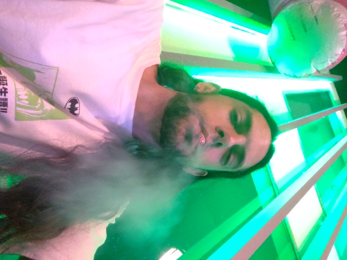 A person wearing a white t-shirt with a Batman logo exhales smoke. The individual has long, dark hair and a beard, and appears to be indoors with green lighting. A glowing green, possibly inflatable, object is visible in the upper right corner. A series of illuminated, stacked shelves are in the background, adding to the vibrant, green-lit scene.
