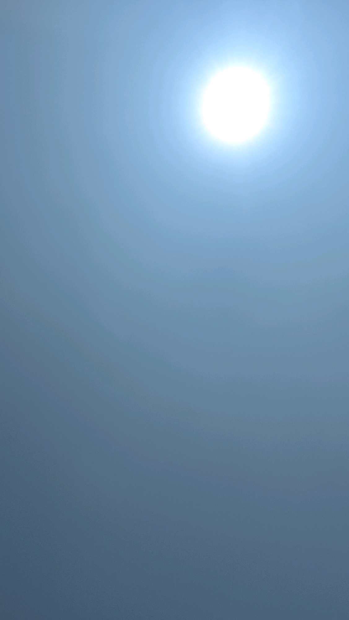 The image shows a bright sun against a pale blue sky. There are no people or man-made objects visible. The image is a photograph of the sky, likely taken in an outdoor setting. The specific location in Slawi, Indonesia, cannot be determined from the image alone.