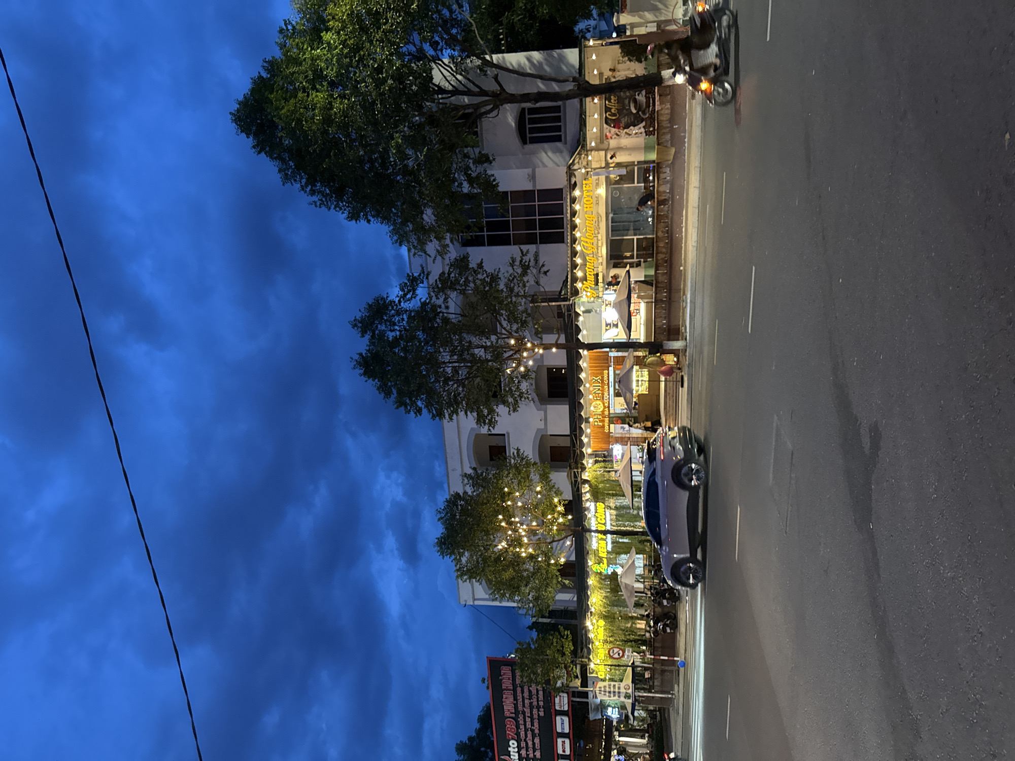This image depicts a street scene in Nha Trang, Vietnam, at dusk or early evening. The sky is a deep blue with dramatic, cloudy formations. A dark power line stretches across the upper portion of the frame.
In the mid-ground, a multi-story building with white walls and dark-paned windows is illuminated by string lights. A sign above a ground-floor establishment reads "PHUONG HOANG HOTEL" and "Coffee". Several trees with lights wrapped around their branches are positioned in front of the building. Outdoor seating with umbrellas is visible under awnings.
A light-colored SUV is driving on the asphalt road in the foreground, with white lane markings visible. To the right, a person on a motorcycle is also on the road, with its headlight illuminated. The road itself shows signs of wear.
On the left side of the image, a large billboard with Vietnamese text and logos is partially visible. Several motorcycles are parked in front of the buildings. The overall impression is a bustling urban environment at the transition from day to night.