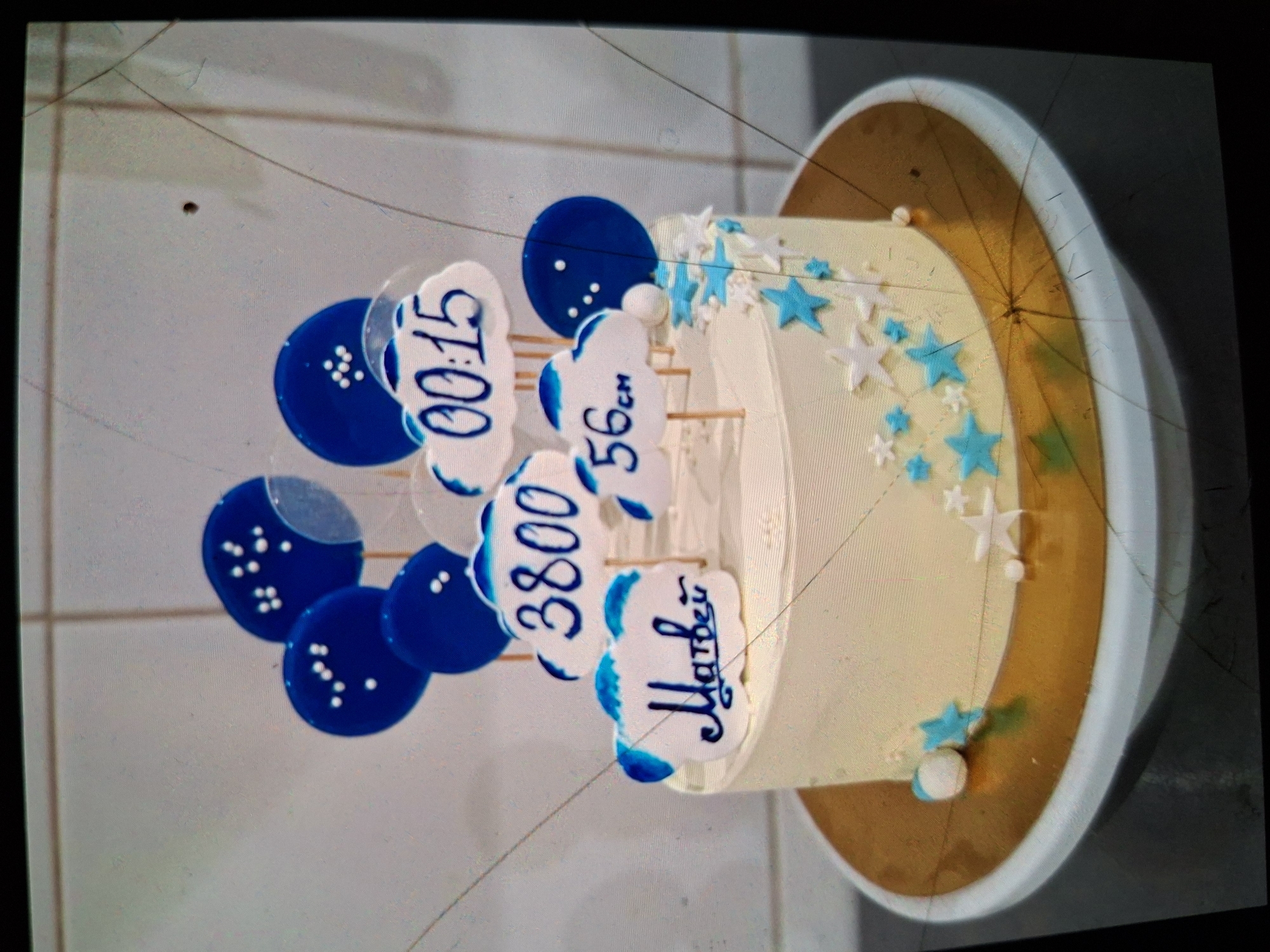 A frosted cake decorated with blue and white cloud-shaped toppers, round blue balloons, and star-shaped sprinkles is displayed on a gold cake board. The cloud toppers feature writing and numbers, including "Матвей" (Matvey), "3800", "56см" (56 cm), and "00:15". The cake itself is decorated with blue and white stars. The background appears to be a wall with a grid pattern, possibly tiled. The image is slightly distorted and has visible cracks, suggesting it may be a photograph of a screen or a damaged print. The location of Medvezhyi Ozyora, Russia is not evident in the visual information provided.