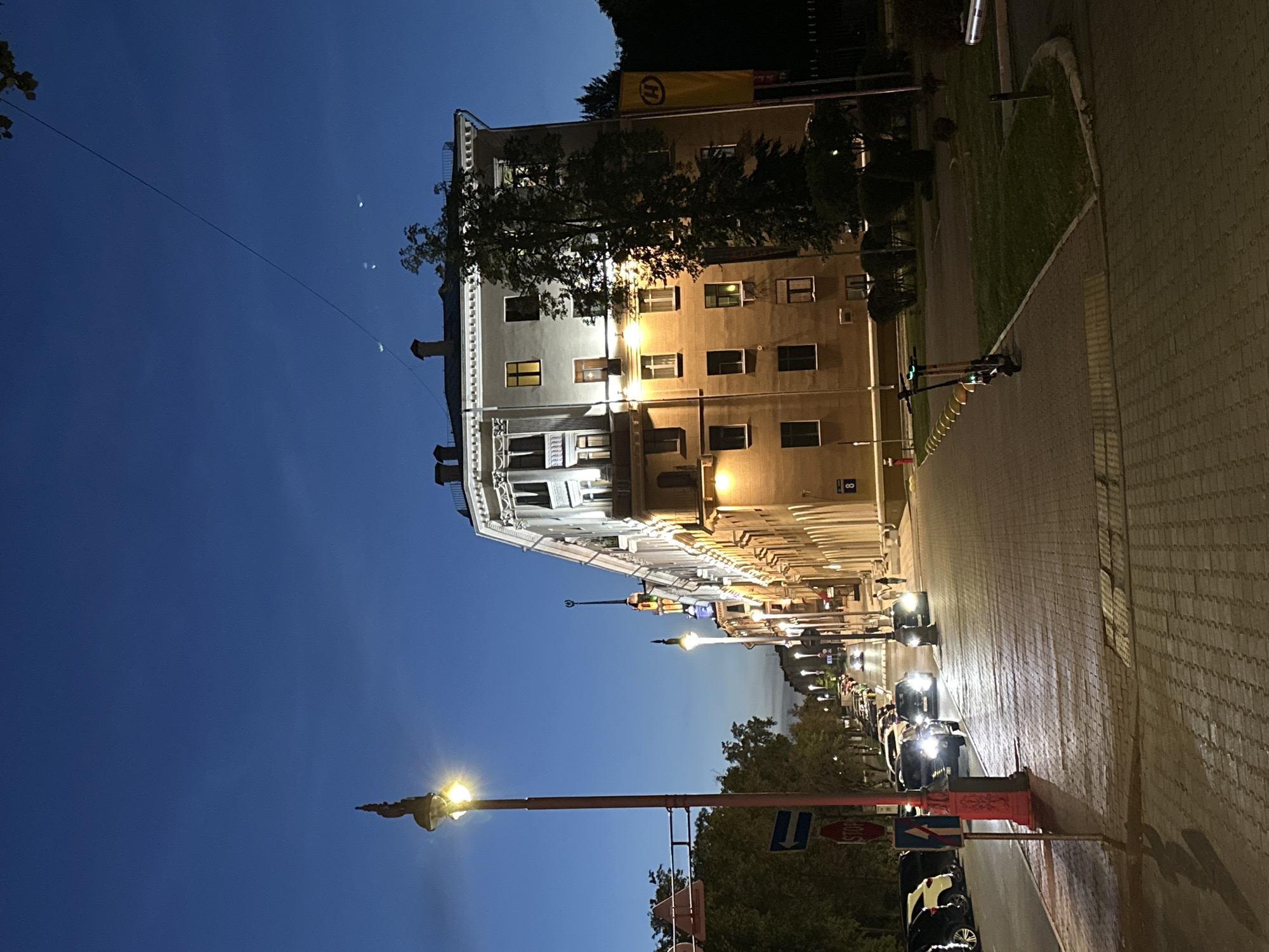 An outdoor, street-level, night-time, vertical, medium shot captures a perspective looking up a cobblestone street in Minsk, Belarus. The street is lined with parked cars on the left and a large, multi-story building on the right. The building is illuminated by warm artificial light, highlighting its architectural features. The sky above is a deep twilight blue.
In the foreground, a red lamppost stands on the left, its light illuminating the street and a stop sign. A blue directional arrow sign is also visible. Further down the street, several cars are driving, their headlights on. Two electric scooters are parked on the sidewalk on the right side of the image. A yellow banner with a black "H" logo is visible on the building's facade.
The scene depicts a quiet urban environment at dusk or early night. The lighting suggests the transition from day to night, with artificial lights beginning to dominate the scene. The presence of cars and scooters indicates typical urban activity. The architecture of the building suggests a historical or governmental structure, common in European cities. The overall atmosphere is calm and serene.