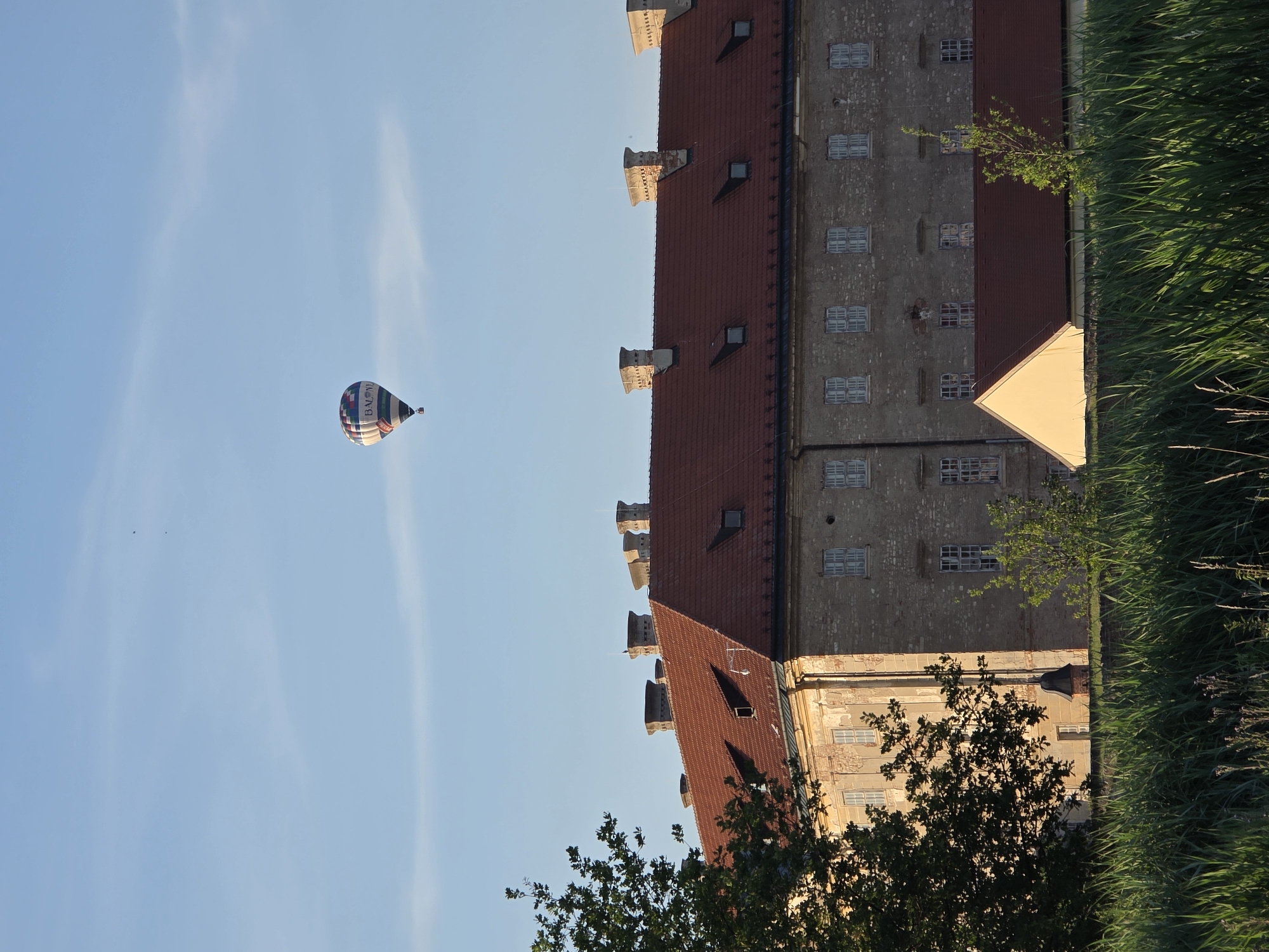 A hot air balloon with red, white, blue, and yellow panels, and the word "BALON" visible, floats in a clear blue sky above a large, multi-story building with a red tiled roof. The building, constructed of light-colored brick, has numerous windows and several prominent chimneys. In the foreground, tall green grass and a portion of a tree are visible. The scene appears to be outdoors during daylight.