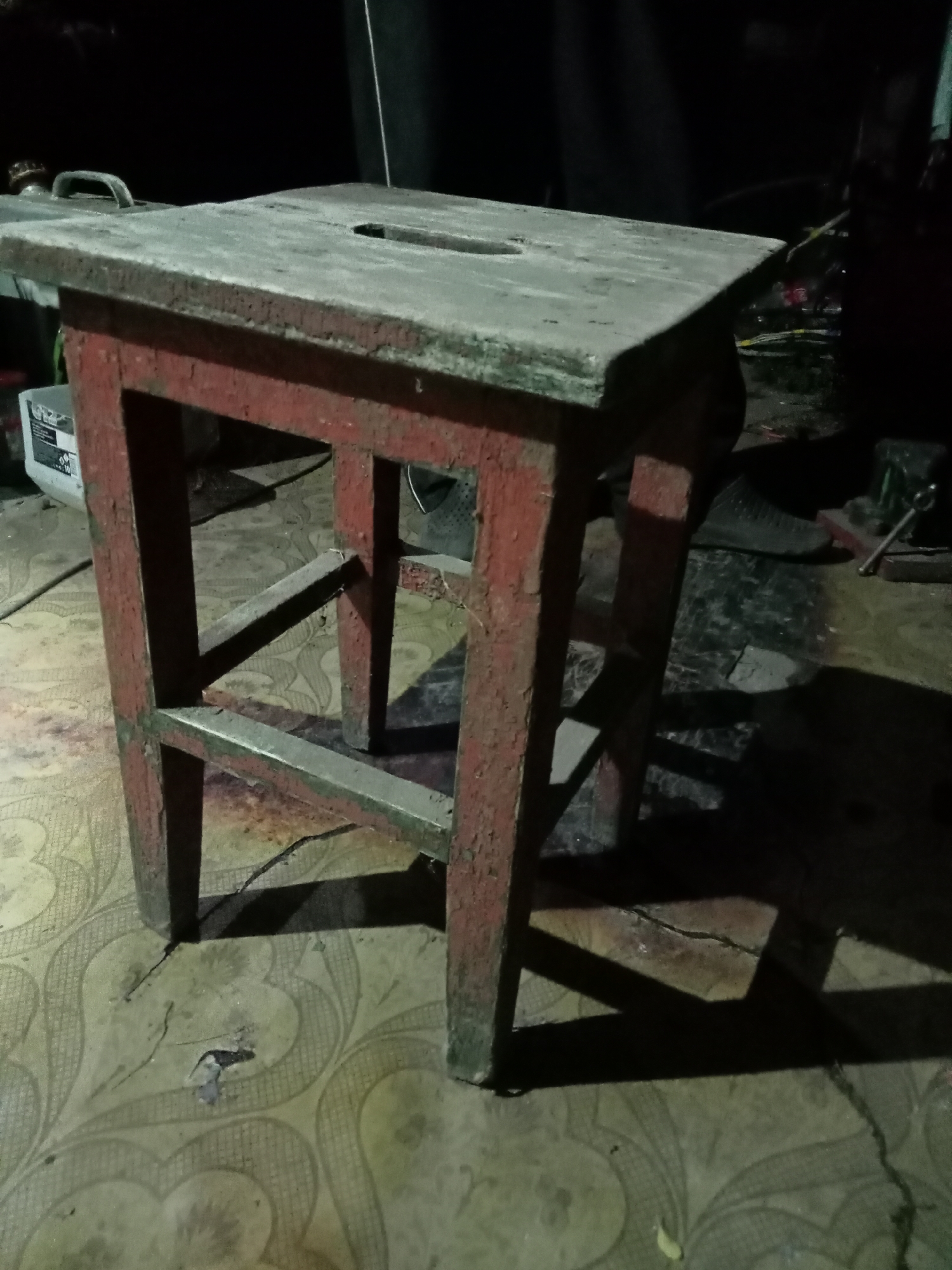 A low-angle, close-up shot focuses on a weathered red stool with a grey, rectangular top. The stool is positioned on a patterned floor, likely linoleum or tile, which appears worn and cracked in places. The lighting is dim, casting long shadows from the stool onto the floor. To the left of the stool, a white plastic container with some black lettering is visible, and behind the stool, the background is largely obscured by darkness, with hints of other objects and surfaces. The overall impression is one of an indoor, possibly workshop or storage area, setting, with the stool being the primary subject.