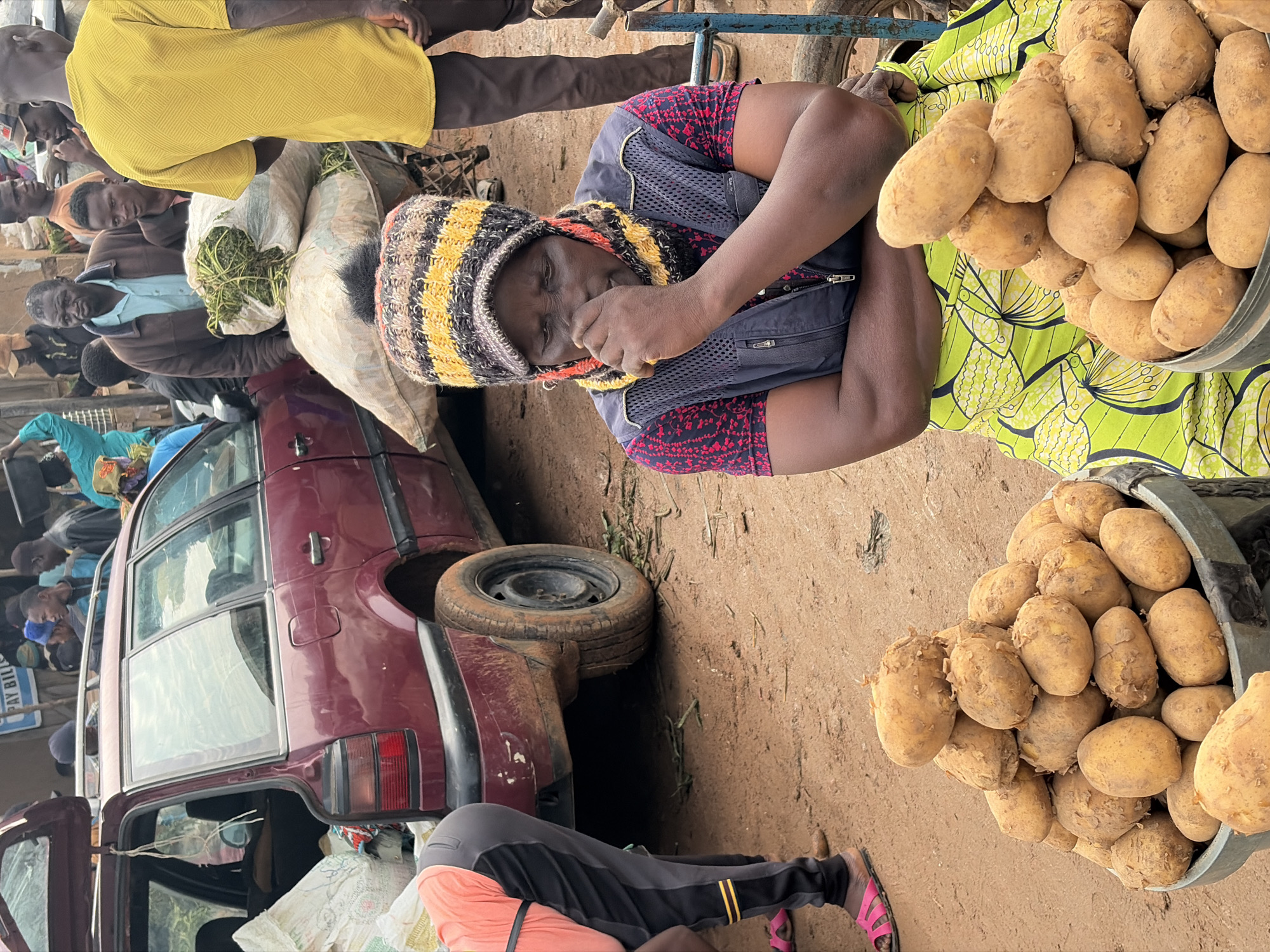 The image depicts an outdoor market scene in Gura Top, Nigeria. In the foreground, a woman wearing a striped beanie, a dark vest over a red and black patterned shirt, and a vibrant green patterned skirt sits with her arms crossed and her hand touching her nose. In front of her are two large containers overflowing with potatoes. To the left, a person wearing an orange shirt and dark pants with yellow stripes is bending down, with pink sandals visible. Behind the woman, a dark red station wagon is parked with its trunk open, revealing bags inside. A wheelbarrow loaded with green leafy vegetables is also visible, with several men standing around it. The ground is dirt and appears to be a market thoroughfare, with debris scattered around. Other individuals and market stalls are discernible in the background, suggesting a busy commercial environment.