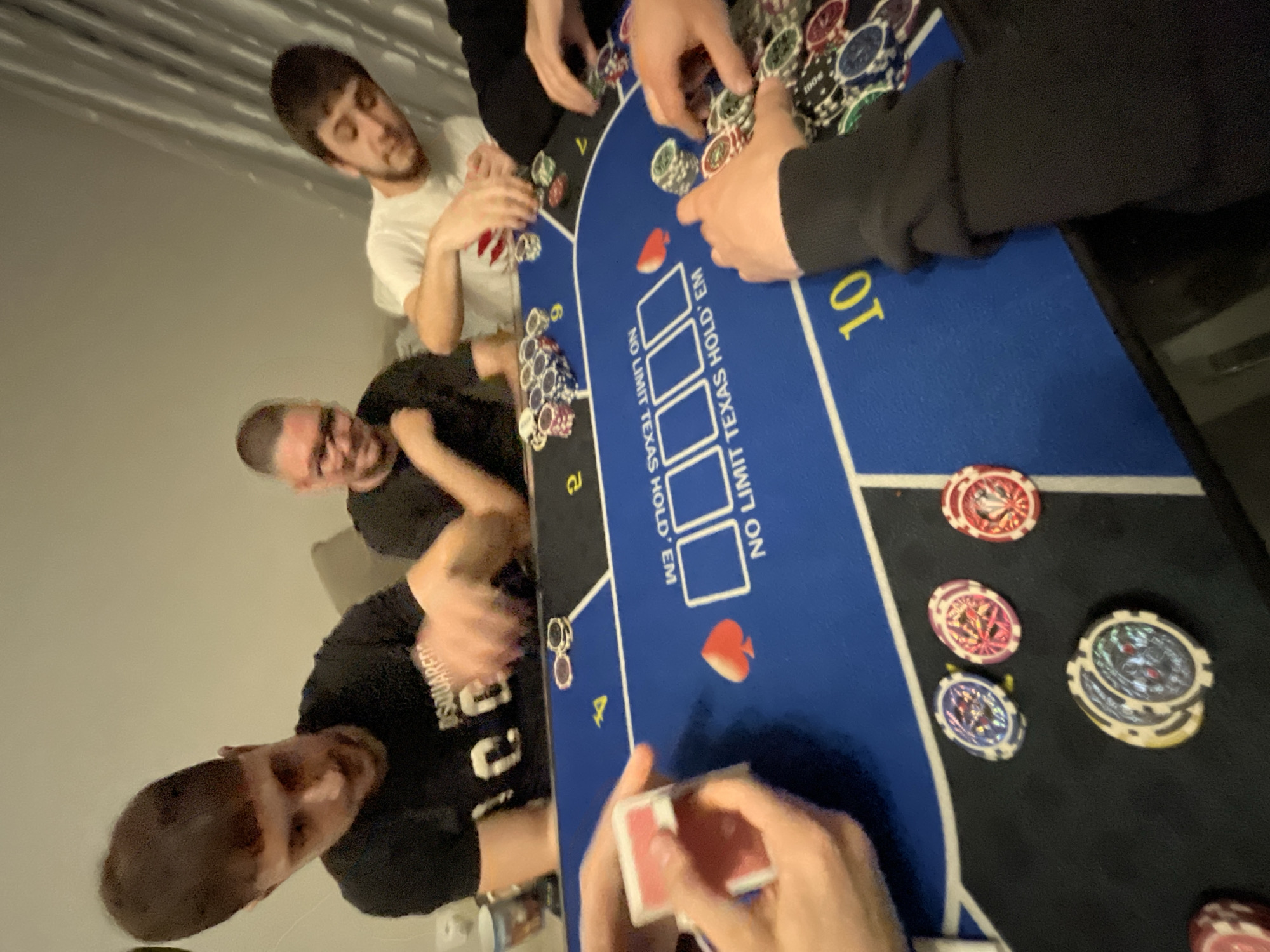 This image depicts a casual game of No Limit Texas Hold'em poker, likely taking place indoors at a private residence in Burgas, Bulgaria. At least five individuals are actively engaged around a blue and black poker table.
Four men are visible: one in a white t-shirt, another in a black t-shirt with "SQUARED" branding, and a third wearing a black long-sleeved shirt. A fourth person's hands are visible on the bottom right, holding a deck of red playing cards. The players are focused on the game, with hands on or near stacks of poker chips. The man in the black "SQUARED" t-shirt exhibits a slight smile.
The central blue section of the poker table is marked with the text "NO LIMIT TEXAS HOLD' EM" and features four empty rectangular boxes, presumably for community cards. A red spade symbol is also printed on this section. The black felt area of the table is covered with numerous poker chips of various colors including red, blue, green, white, black, yellow, and purple, some with designs suggesting denominations like "100". Numbered positions "7", "8", and "10" are visible along the table's edge. The ambient indoor lighting suggests the game is taking place in the evening or at night. The scene conveys an atmosphere of focused recreational activity.