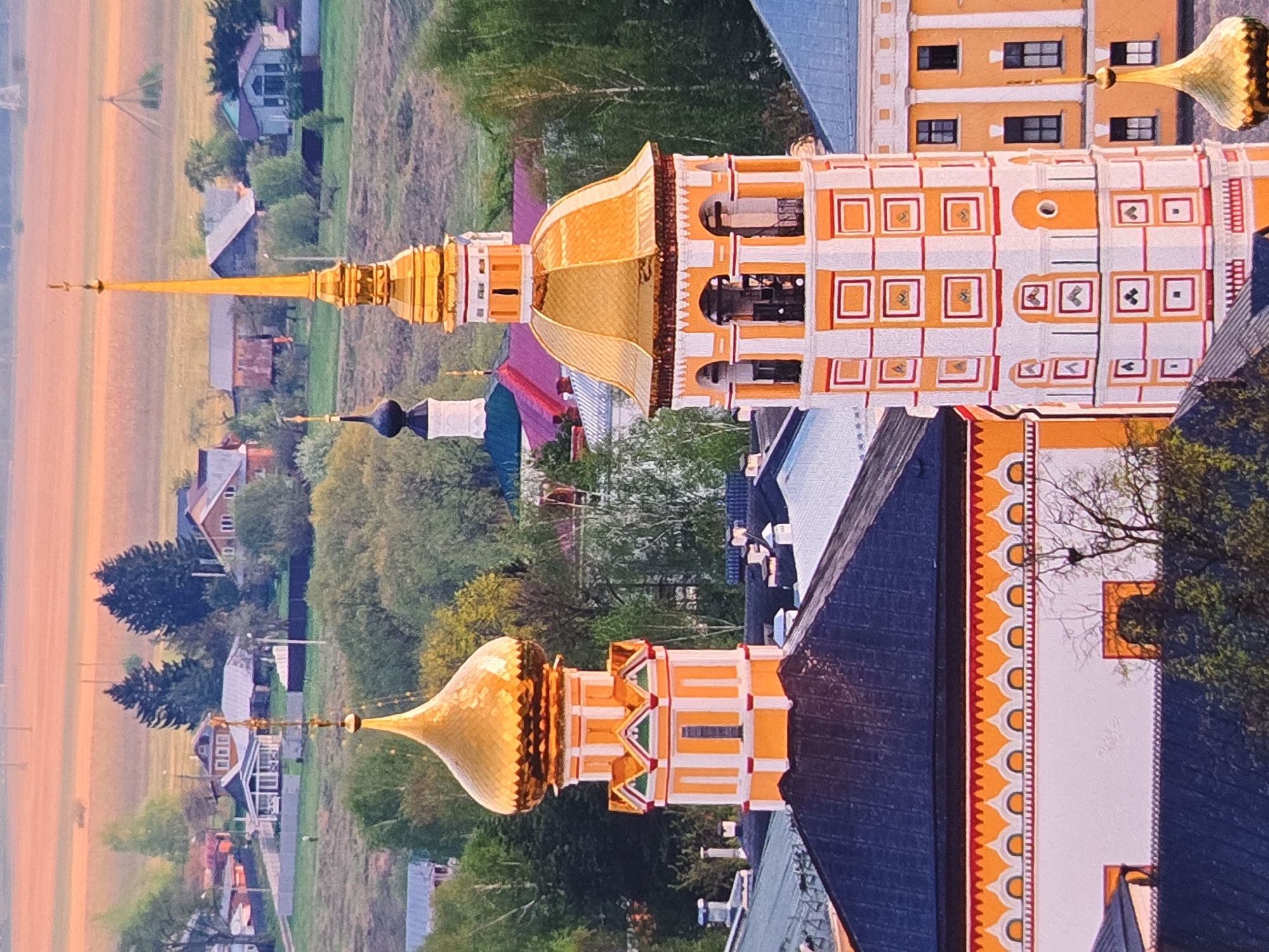 This is a vertical, medium-angle, eye-level shot of the Assumption Cathedral in Vladimir, Russia, captured during what appears to be late afternoon or early evening. The sky is a soft, warm gradient of orange and pink, suggesting a sunset. The overall scene is peaceful and serene, with the golden domes of the cathedral glinting in the soft light.
The primary subject is the impressive architecture of the Assumption Cathedral, with its distinctive golden domes and ornate detailing. The cathedral is painted in bright white and orange stripes, with decorative patterns and small square windows. The golden spire of one dome is particularly prominent, reaching towards the sky. In the background, a rural landscape unfolds with scattered houses, some with red roofs, surrounded by lush green trees and fields. A few smaller, darker domes are also visible, possibly belonging to other churches.
The image focuses on the grandeur of the cathedral and its integration into the natural and built environment of the surrounding village. There are no people visible in the shot, and the activity appears to be static, showcasing the timeless beauty of the religious structure. The weathered textures of the buildings and the soft, diffused light contribute to a sense of history and tranquility. The location is identifiable as Vladimir, Russia. There is no visible text within the image.