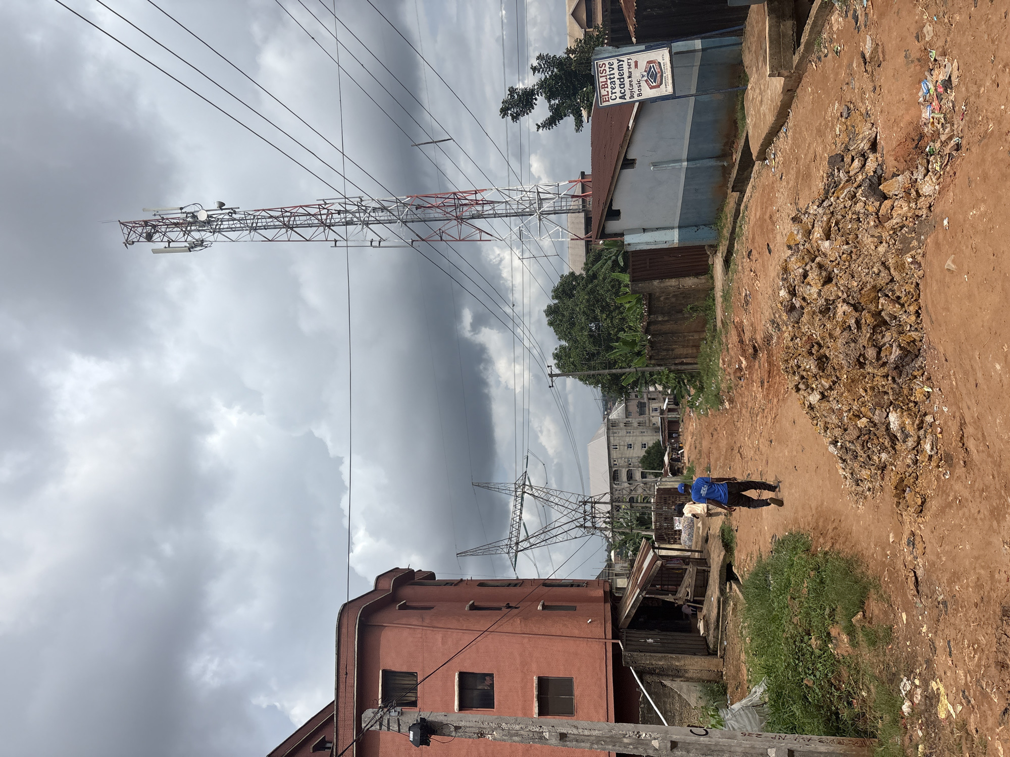 This media file depicts a scene in Nkpor, Nigeria. The image is oriented vertically, showing a street with a large telecommunications tower dominating the left side. The tower is red and white, with numerous cables running from it across the sky. In the background, there are several buildings, including a salmon-colored building with multiple windows and a blue and grey building with a sign that reads "EL-BLISS Creative Academy Day Care. Nursery Basic".
The ground is a dirt path, with piles of rocks and debris, including plastic waste, scattered in places. Two individuals are walking on the path, one wearing a blue shirt and dark pants, the other partially visible in a blue shirt and dark pants as well. There is a small, open-sided structure with a corrugated metal roof to the right of the salmon-colored building, and some vegetation. The sky is overcast with dark clouds, suggesting a cloudy or potentially rainy day. The presence of the telecommunications tower and the style of the buildings suggest an urban or semi-urban environment. The overall impression is one of a functional, everyday street scene in a developing area, with a mix of infrastructure and some signs of litter and disrepair.