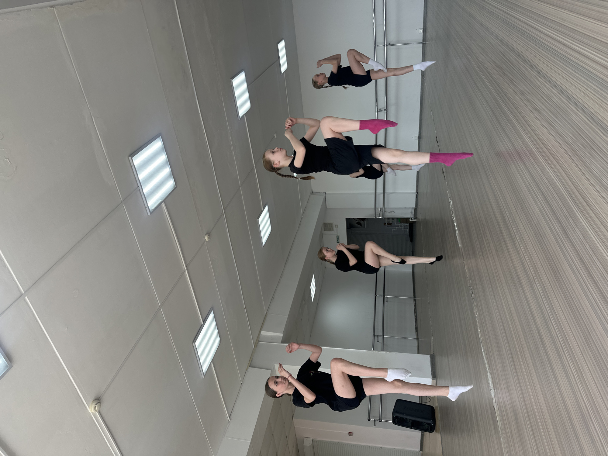 The image shows four young girls in a dance studio. They are all wearing black outfits and socks. The girls are lying on the floor, seemingly practicing dance poses. The studio has a light-colored floor and walls. There are rectangular ceiling lights. Ballet barres are along the wall. This may be in Votkinsk, Russia, due to the source.
