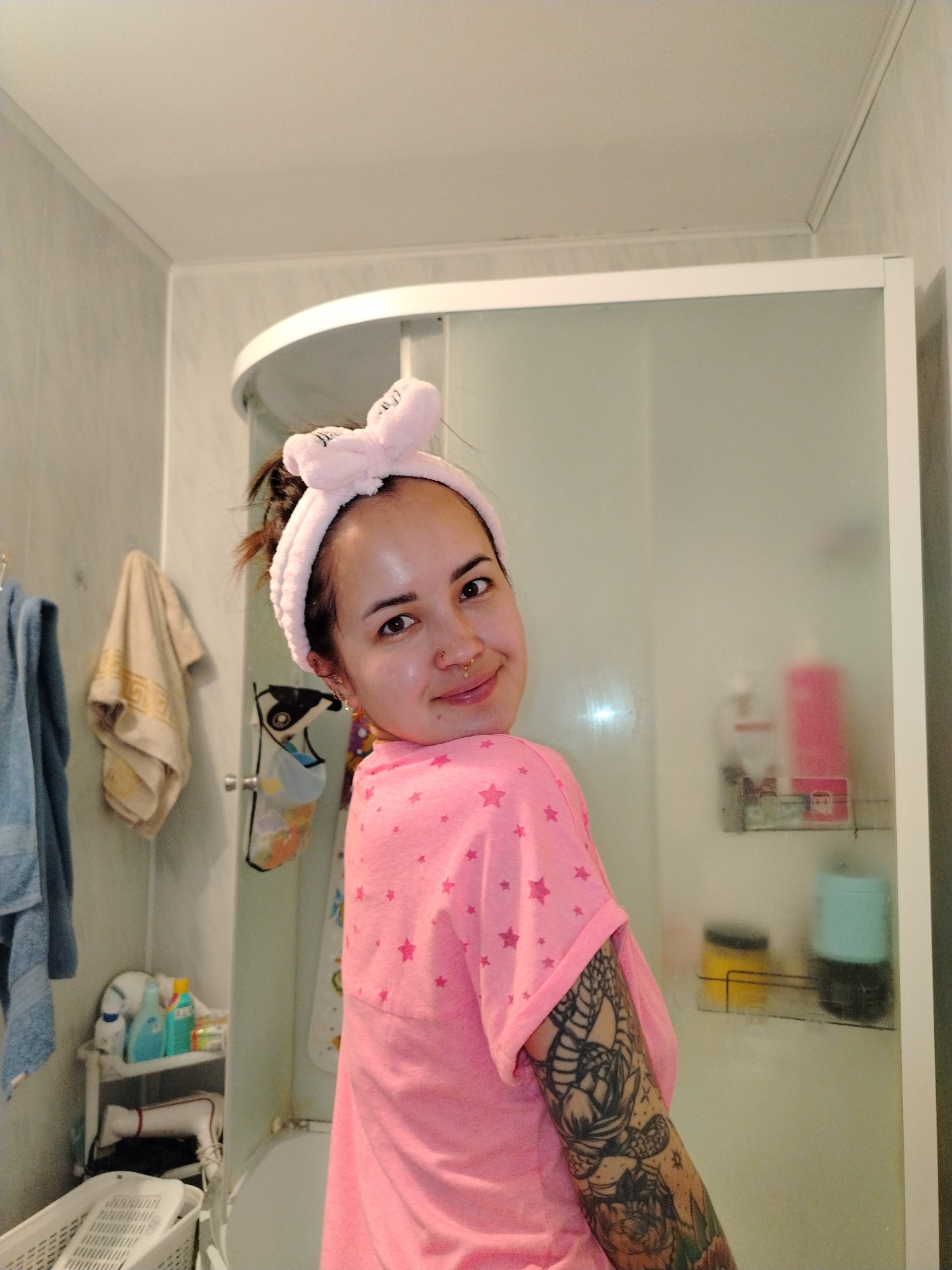 A young woman with a pink headband and matching pink shirt with star patterns is in a bathroom. The bathroom features a shower stall with frosted glass doors and a white frame. Inside the shower, there are several bottles and containers on a wire shelf. To the left of the shower, there are towels hanging on a rack and a small shelf unit with various toiletries. A laundry basket is visible at the bottom of the frame. The woman has tattoos on her left arm and is looking back at the camera with a slight smile. The bathroom walls are a light gray color, and the ceiling is white.
The location is not possible to know based on the image.