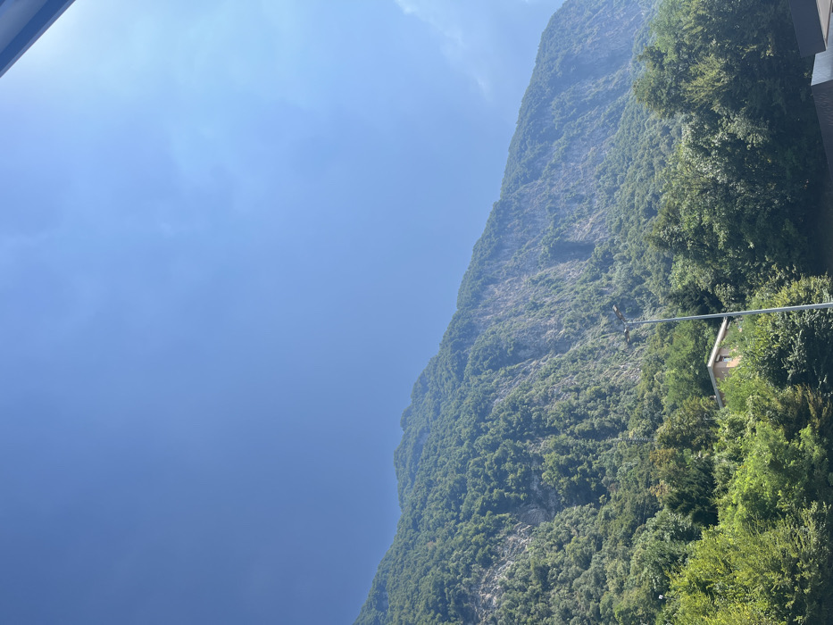 The image captures a steep, heavily forested mountain slope under a clear blue sky, characteristic of the mountainous terrain immediately surrounding Mendrisio, Ticino, Switzerland. The sky, occupying the upper half of the frame, is a vibrant blue with faint, diffuse white cloud formations visible in the upper right quadrant. The lower half of the scene is dominated by a large mountain or hill, densely covered with green deciduous and coniferous trees. Prominent grey rock faces and sections of sheer cliff are visible throughout the tree cover, particularly on the central and right portions of the slope. In the lower-central foreground, a grey, cylindrical lamppost with a light fixture extends vertically. To the left of the lamppost, a portion of a light-tan or beige building, featuring a sloped brown roof, is partially visible nestled within dense green foliage. A dark, flat, angular surface is partially visible in the extreme lower right corner, indicating a nearby man-made structure or ground feature. The scene is illuminated by bright daylight. No individuals are visible.