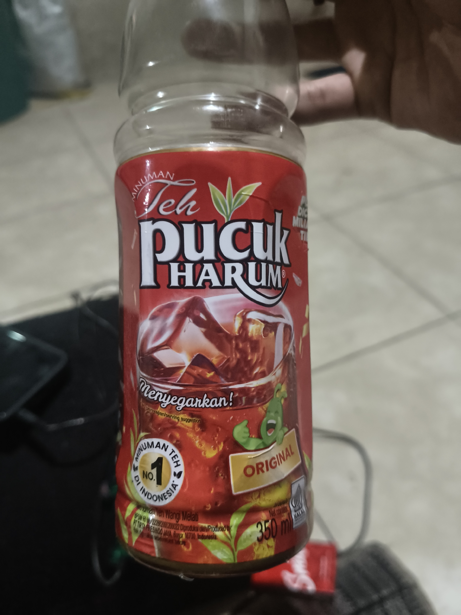 In the city of Ulunipa, Indonesia, an outdoor scene is captured, focusing on a plastic bottle of "Teh Pucuk Harum" tea. The bottle, predominantly red with white lettering, displays the brand's logo and imagery of iced tea. The label also features text in Indonesian, including "MINUMAN", "Teh Pucuk Harum", "Menyegarkan!", "ORIGINAL", and "350 ml". A small green cartoon character with its thumb up is visible on the label, positioned next to the word "ORIGINAL". The bottle is held by a person, whose hand is partially visible in the upper right corner of the frame, indicating a casual, handheld presentation of the product. The background is blurred but suggests an indoor or semi-outdoor environment with tiled flooring and indistinct objects. The lighting appears to be ambient, with the primary focus on the product.