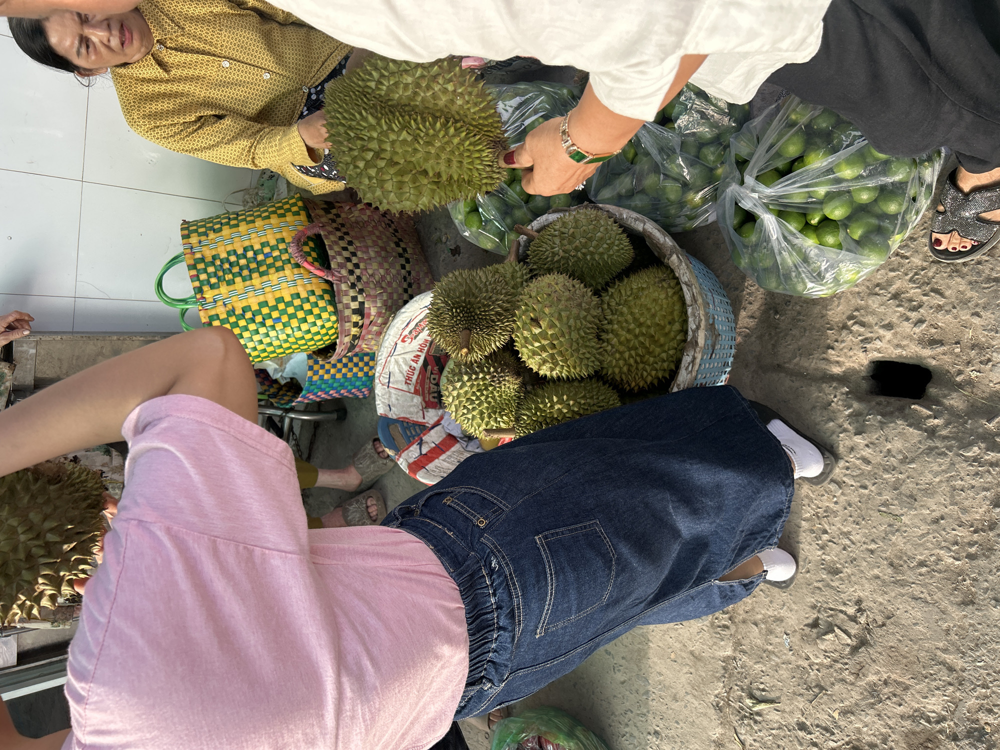 The image depicts an outdoor market scene in An Hữu, Vietnam, likely near a street or roadside given the rough, sandy ground. In the foreground, a person wearing a pink t-shirt and a dark denim skirt holds a durian fruit. To the right, another person in a white shirt is selecting a durian from a basket. A woman in a yellow patterned shirt is seated behind a display of woven baskets, one of which contains durians. Several durian fruits are piled in a blue woven basket. Bags of green limes are also visible. The overall atmosphere suggests a vibrant fruit market.