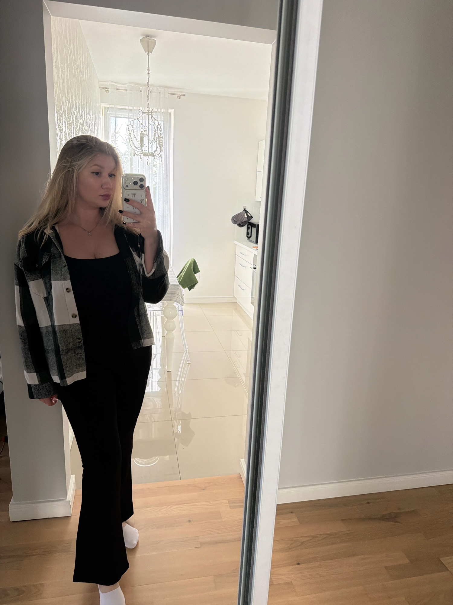 In Koszalin, Poland, a young woman is captured in a mirror selfie taken in what appears to be a modern apartment. She stands in a hallway, leaning against a light gray wall. She is wearing a black sleeveless top, black flared pants, white socks, and an open black and white plaid shirt. Her blonde hair is down, and she is holding a smartphone to take the picture. The reflection in the mirror reveals a well-lit room with a chandelier, a window with sheer curtains, and a glimpse of a kitchen area with white cabinets and a microwave. The flooring transitions from light-colored tiles in the background to wooden planks in the foreground.