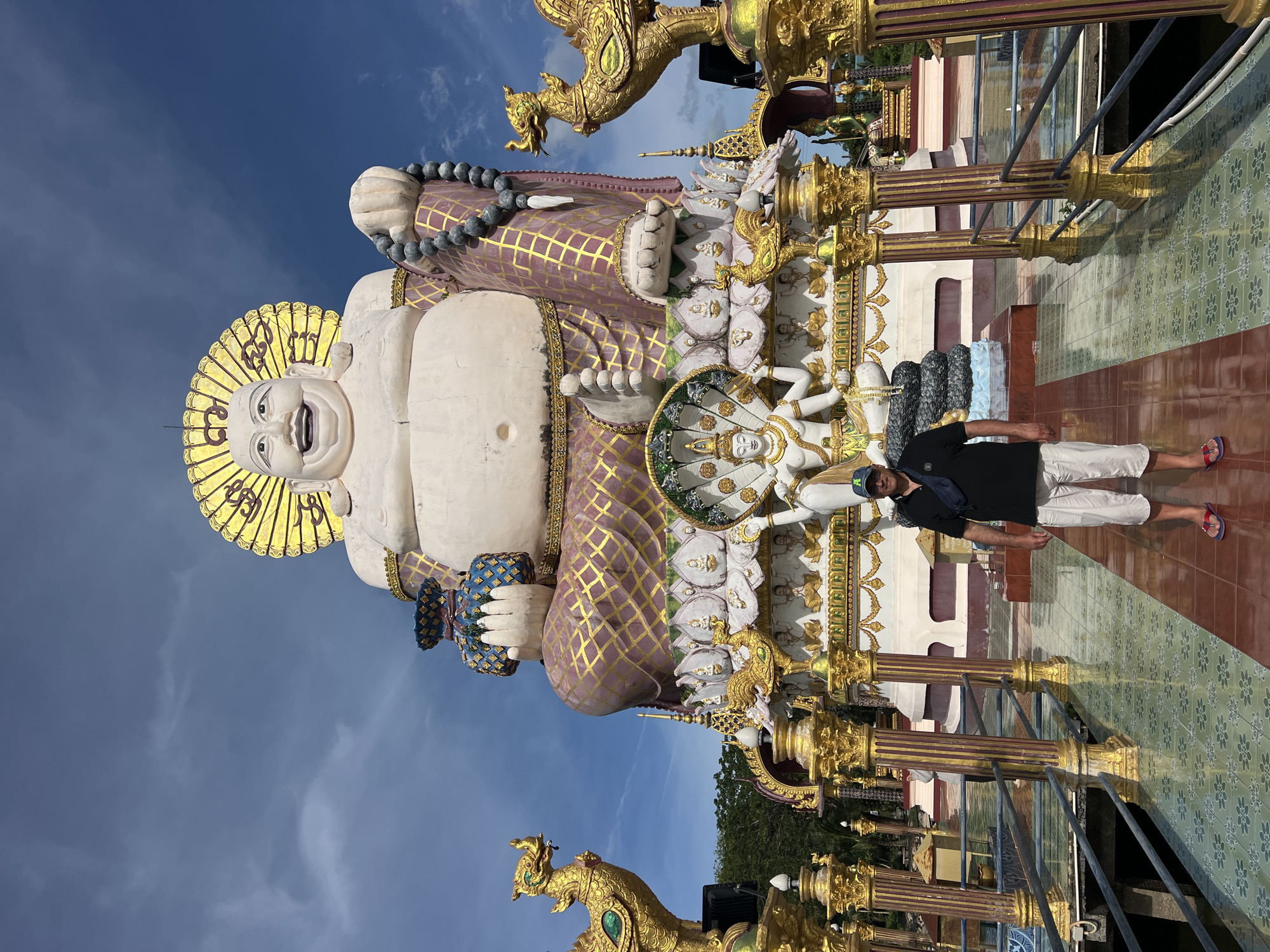 A full, slightly low-angle shot shows a man standing in front of a large, seated Buddha statue on a sunny day. The man, who is wearing a black t-shirt, white shorts, a blue baseball cap, and red flip-flops, stands facing the camera. He is positioned on a walkway that leads towards the statue. The walkway is adorned with ornate, gold-colored pillars and railings. The colossal Buddha statue is depicted with a cheerful expression, holding a beaded necklace in its right hand and a blue and gold patterned bag in its left. The statue is adorned with a golden halo and intricate purple and gold patterned robes. Below the main Buddha statue, smaller figures and decorative elements are visible, including what appears to be a multi-headed Naga serpent. The scene is set against a bright blue sky with scattered white clouds, and lush green trees are visible in the background, suggesting a tropical location. The overall impression is one of grandeur and cultural significance, likely a tourist attraction. This statue is the Big Buddha of Koh Samui, located in the Wat Phra Yai temple complex in the Ko Samui District of Thailand.