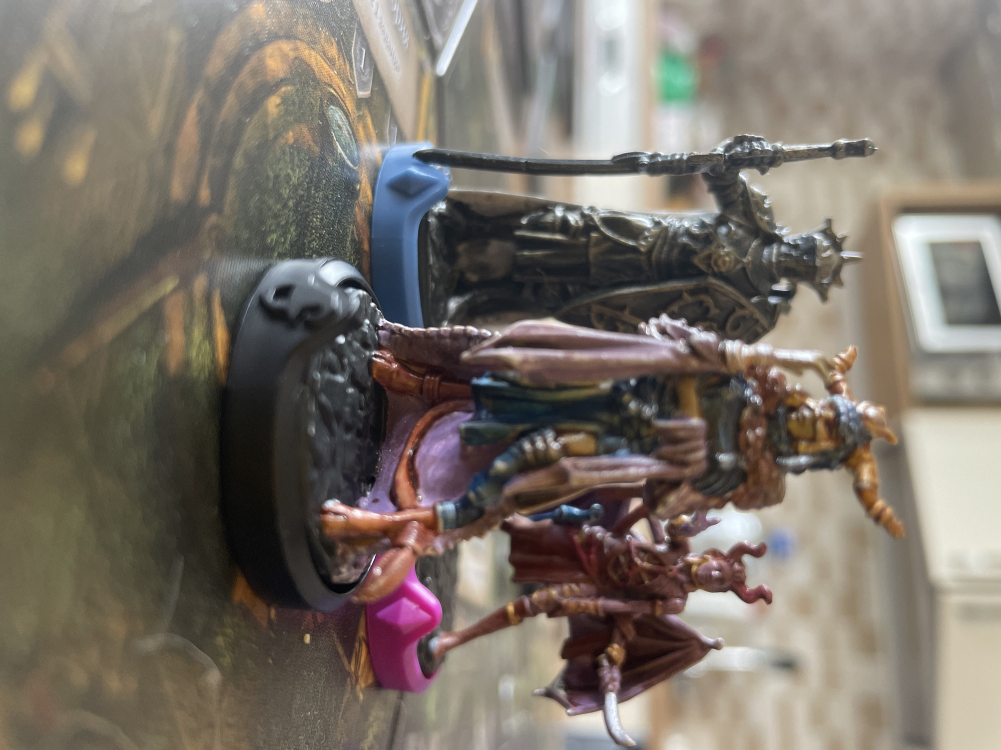 The image shows three miniature figurines, likely for a tabletop game, positioned on a game board.
The figurines are arranged in a row from left to right. The figure on the left is atop a circular base. Next, a miniature with a purple base and the third is on a blue base.
The background depicts a game board with artwork, predominantly brown and green in color. There is nothing in the photograph to suggest this image was taken in the city of Budva, Montenegro.
