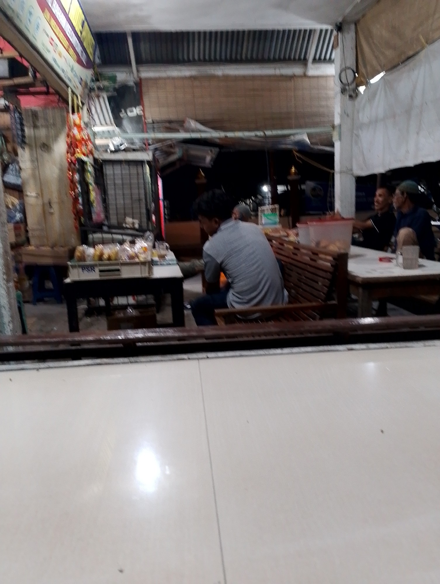In the city of Bireuenmeunasahreuleut, Indonesia, an outdoor food stall is depicted. A young man with dark hair, wearing a grey t-shirt and dark pants, is seated facing away from the camera, looking at a table laden with food items and drinks. He is on a wooden bench, possibly part of a seating area. To the right, two other men are seated at a separate table. One man is wearing a dark t-shirt and appears to be talking and smiling, while the other, wearing a blue shirt, has his back partially to the camera. In the foreground, a white tiled floor reflects light. The stall is open, with various goods displayed on shelves and racks in the background, suggesting a bustling commercial area.