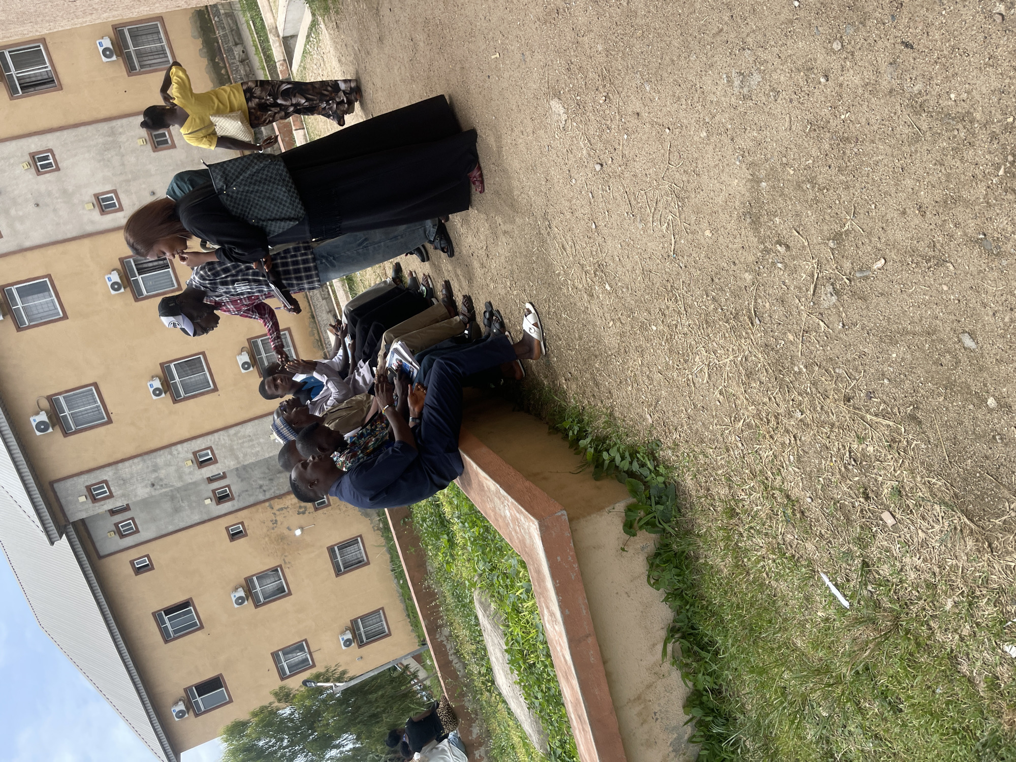 In Jos, Nigeria, on a bright day, a group of individuals are gathered outside a three-story building with a pale yellow facade and a silver metal roof. The building features numerous windows with white frames, some equipped with air conditioning units. The ground is a mixture of dirt and sparse dry grass, with a low, concrete retaining wall covered in green foliage.
Several men are seated on the retaining wall, facing left. One man in a dark blue outfit is looking down at papers in his lap. Next to him, another man in a colorful patterned shirt is also seated. Further down the wall, a group of men are seated together, some looking at phones, others engaged in conversation. One man is wearing a white and blue patterned cap.
Standing and interacting are two individuals. A man in a red and black plaid shirt and blue jeans is holding a black object, possibly a tablet or book. A woman in a long black dress and a patterned bag is speaking with him. Another woman in a bright yellow top and patterned pants is walking away, her back to the camera, carrying a white bag. A young person in a black shirt is also partially visible near the standing individuals.
The overall scene suggests an informal gathering or meeting taking place outdoors, with the building and the surrounding environment providing the backdrop.