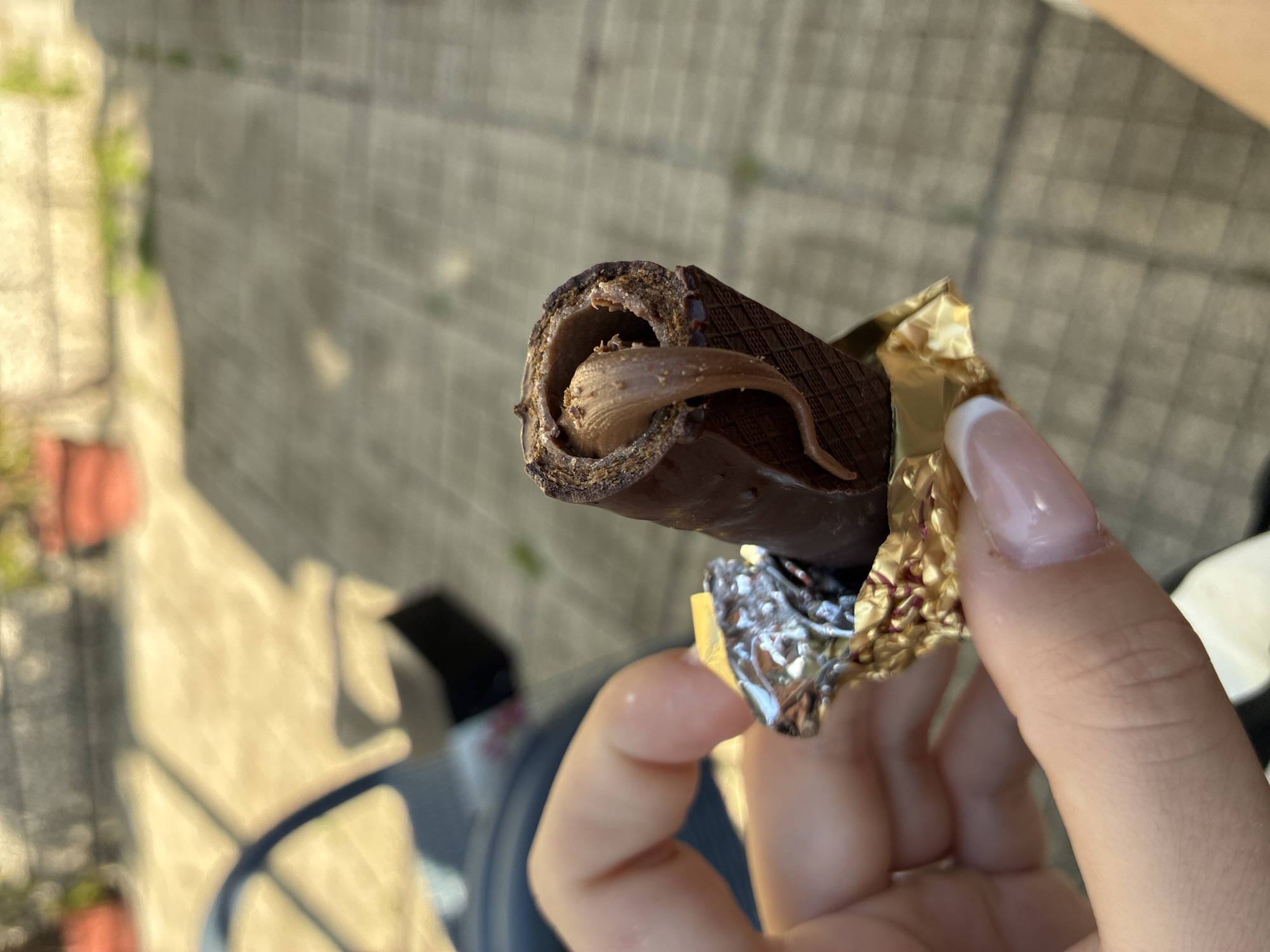 The image displays a human hand holding a partially unwrapped chocolate-covered wafer roll. The hand, featuring manicured nails with a white tip design, grasps the gold-colored foil wrapper. The chocolate roll exhibits a textured, grid-like pattern on its exterior. A lighter brown, creamy filling is visible within the roll, with a distinct, irregular portion of the filling protruding and curving downwards from the top opening. The background is out of focus, revealing a paved outdoor surface with discernible grid lines and cast shadows, suggesting bright daylight. Indistinct elements in the far background include blurred structures, green foliage, and red-colored objects, possibly potted plants. The scene is located in Klisura, Bulgaria.