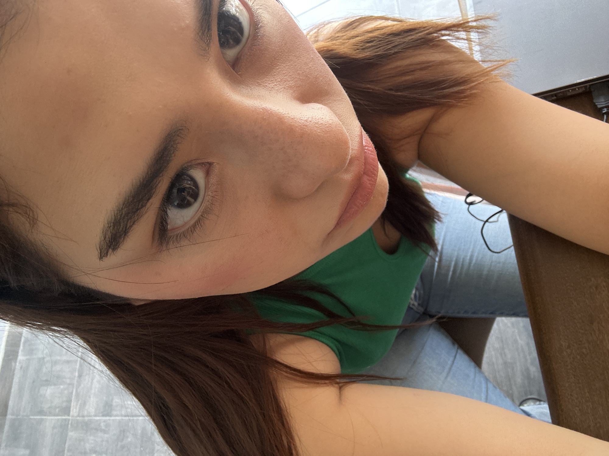 A young woman takes a selfie indoors. Her face is the main focus, angled closely to the camera, showing her brown eyes, dark eyebrows, and light skin with some visible pores and freckles. She has brown hair and wears a green top. Part of her denim jeans and a wooden table are visible in the background. While I can describe the image's elements, I cannot determine the location as Puebla, Mexico, without additional context.