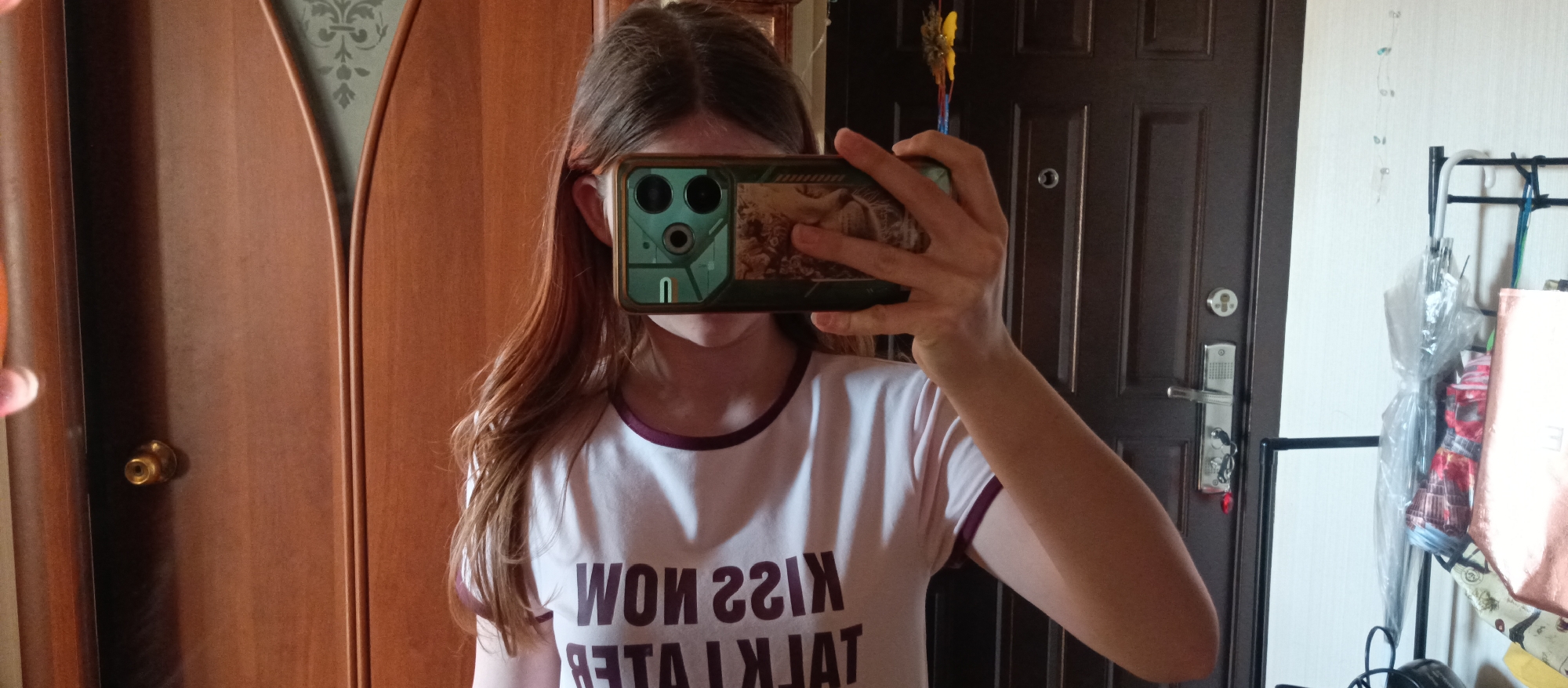 A person is holding a smartphone up to their face, taking a selfie. The smartphone has a green and tan case on it. The person is wearing a white t-shirt with "KISS NOW TALK LATER" printed on it in dark letters. The text is mirrored. The person has long, light brown hair. Behind the person is a closed wooden door with a brass doorknob to the left. To the right is another closed brown door with a silver handle and security lock. Next to that door is a white wall with a black metal rack that holds an umbrella and some bags. The location is Unknown, Unknown.
