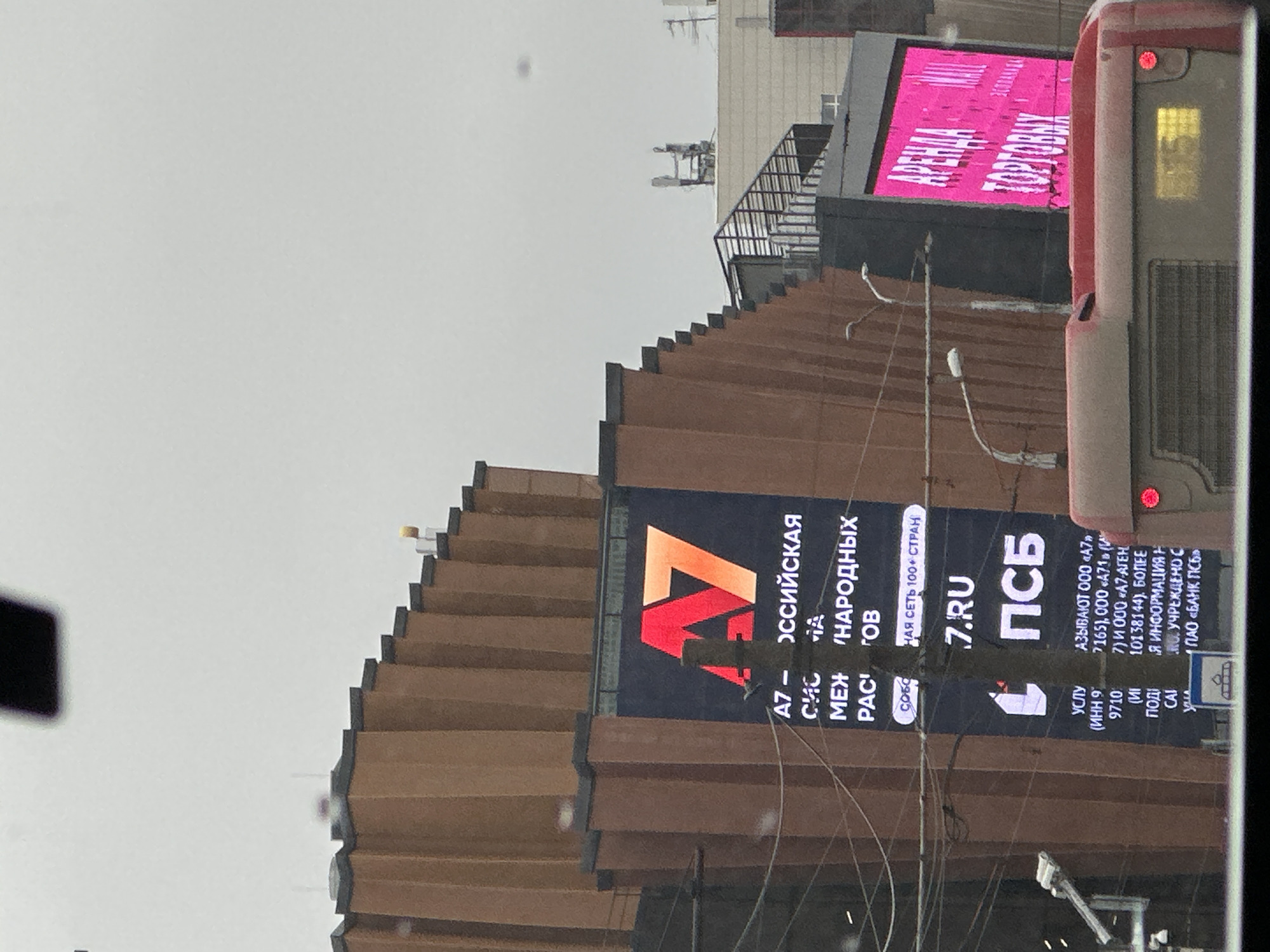 The image captures an urban scene in Perm, Russia. A bus is visible on the right side, with a digital billboard displaying a pink sign reading "АРЕНДА ТОРГОВЫХ". A building with a stepped, brown facade dominates the center, adorned with a large advertisement that includes the "A7" logo, the words "РОССИЙСКАЯ", "НАРОДНЫХ", and the website "7.RU". The abbreviation "ПСБ" is also displayed on the advertisement. Utility poles with wires traverse the scene. The sky is overcast, suggesting cloudy weather.