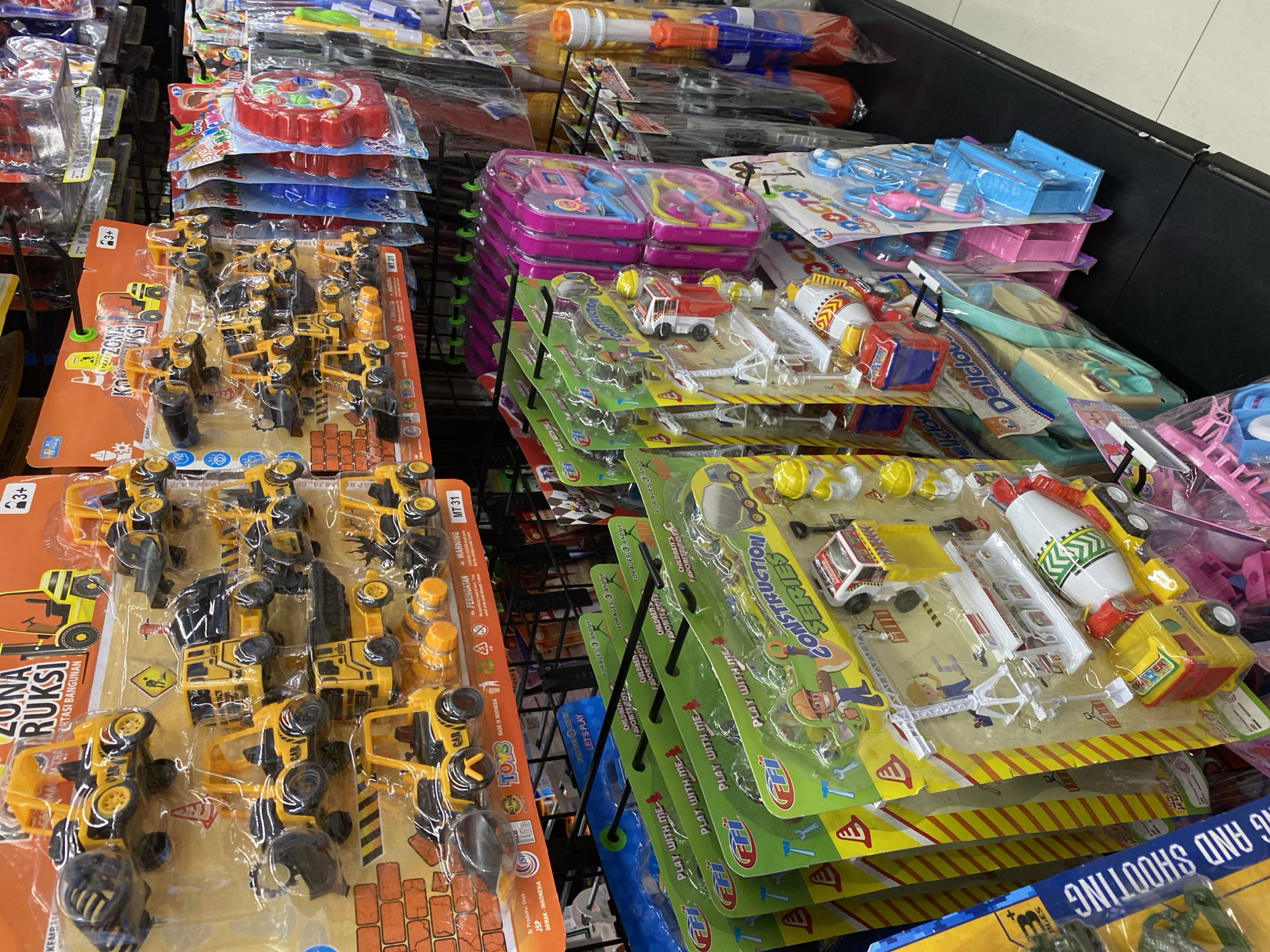The image shows a retail display of children's toys in Banda Aceh, Indonesia. The toys are primarily construction vehicles and medical play sets, packaged in colorful blister packs and hanging from a black metal rack. The construction toys are yellow and black, with visible details like wheels, scoops, and cement mixers. The medical sets are pink and blue, featuring toy stethoscopes, syringes, and doctor's bags. In the background, other packaged toys, including water guns, are visible. The overall environment appears to be a store aisle, with a dark surface in the foreground that could be a shelf or counter. The lighting suggests an indoor setting, possibly a supermarket or toy store. There are no people visible in the image.