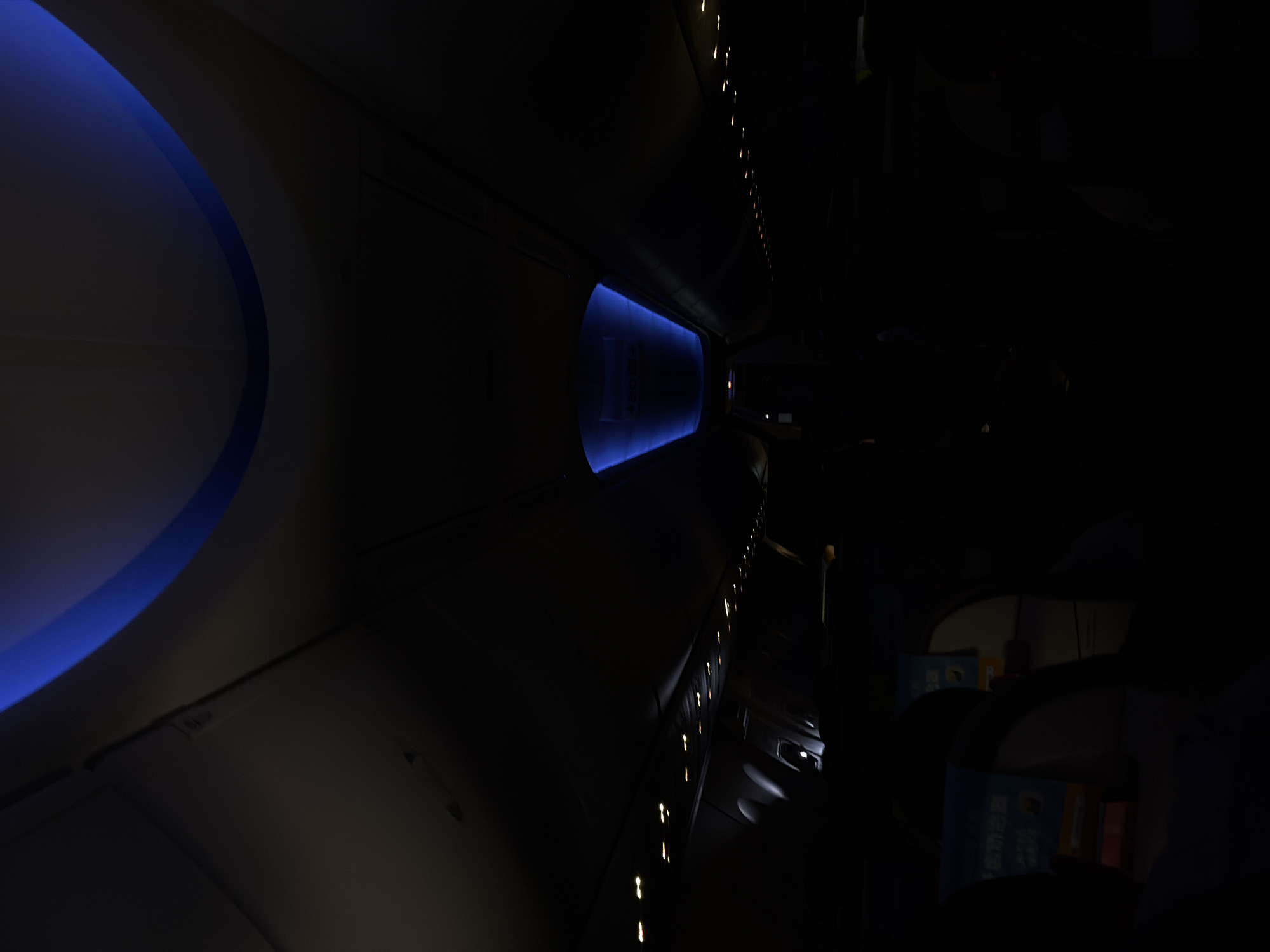 The image shows the interior of an airplane cabin at night. The cabin is dimly lit with blue accent lighting along the ceiling and around the window. Passenger seats are visible along the side, and overhead compartment lights are faintly seen along the top. The scene depicts a flight journey, potentially landing or taking off in Tambon Mai Khao, Thailand.