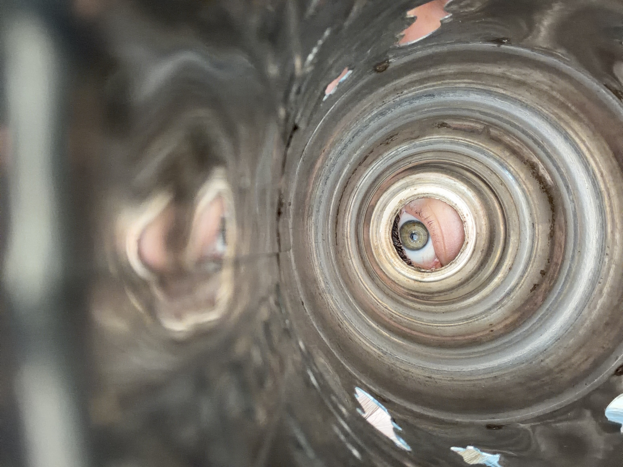 The image is a close-up, distorted, and reflective view looking into a cylindrical object. The object appears to be made of polished metal and has a series of concentric ridges. In the center of the frame, viewed through the cylindrical opening, is a person's eye, looking directly at the camera. The eye is green and appears to be looking through a lens or reflective surface, which distorts its features. The upper portion of the image shows a more abstract, distorted reflection of what appears to be the same person's face, with parts of their mouth and nose visible. The overall impression is one of looking through a narrow aperture at a reflected subject. There is no information in the image to suggest a location in Chaikovsky, Russia.