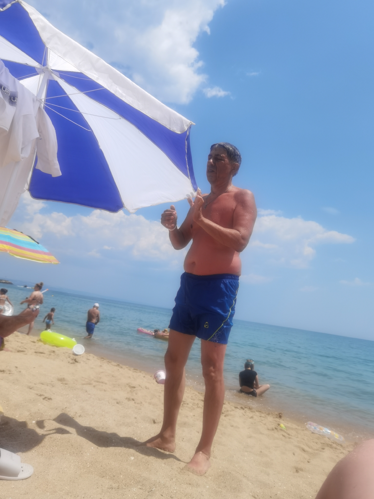 The image captures a sunny day at a beach, likely in Varna, Bulgaria, given the context. In the foreground, a man in blue swim shorts stands facing left, with his right arm raised in a thumbs-up gesture. He is partially shaded by a large blue and white umbrella. Behind him, the sea stretches out, with several other beachgoers visible. Some are in the water, including a man wading and a person on an inflatable raft. On the sandy shore, there are other people, as well as beach accessories like a bright yellow inflatable. The sky is a clear blue with a few scattered white clouds. The overall scene depicts a typical summer day at the beach.
