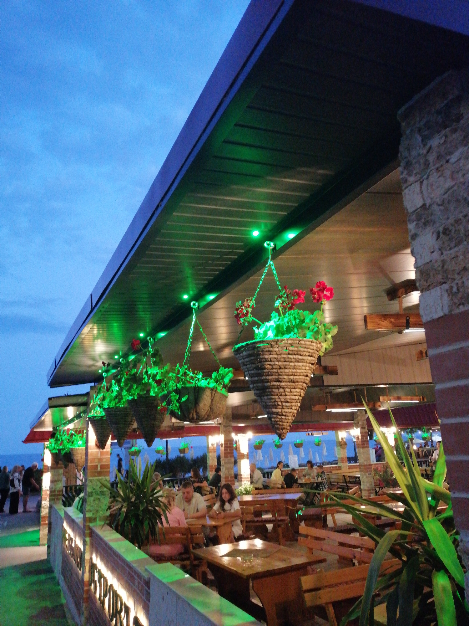 The image depicts an outdoor seating area of a restaurant in Varna, Bulgaria, with a view of the sea and a beach in the background. The sky is a deep blue, indicating dusk or early evening. The restaurant features a covered patio with a dark, sloped roof, illuminated by green LED lights. Several hanging baskets filled with red flowers and green foliage are suspended from the ceiling. The patio is furnished with wooden tables and benches, and a brick wall with illuminated text spelling out "RESTO BAR" is visible in the foreground. A few patrons are seated at tables, some engaged in conversation, while others appear to be dining. In the background, people are walking along a promenade next to the beach, where beach umbrellas are visible. A large green plant is positioned in the right foreground, obscuring some of the seating area. The overall ambiance is relaxed and inviting, suggesting a pleasant evening dining experience by the sea.