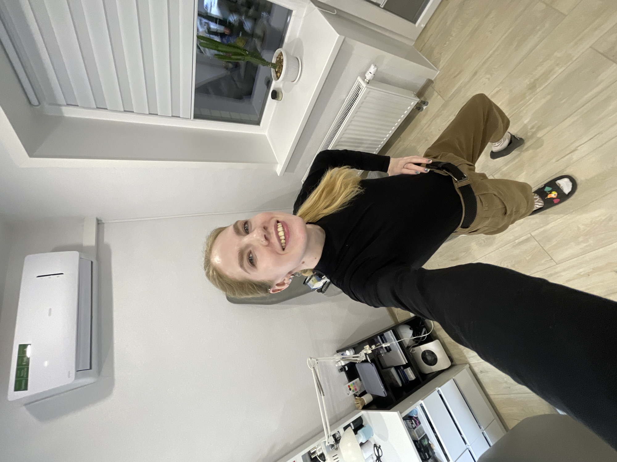 A young, blonde-haired woman is smiling at the camera. She is wearing a black long-sleeved shirt and khaki cargo pants. She is also wearing slide sandals. She is standing inside a room, presumably in Izhevsk, Russia. There is a white air conditioner unit on the wall, a window with blinds, a radiator under the window, and a white desk with various objects on it. The floor is tiled.
