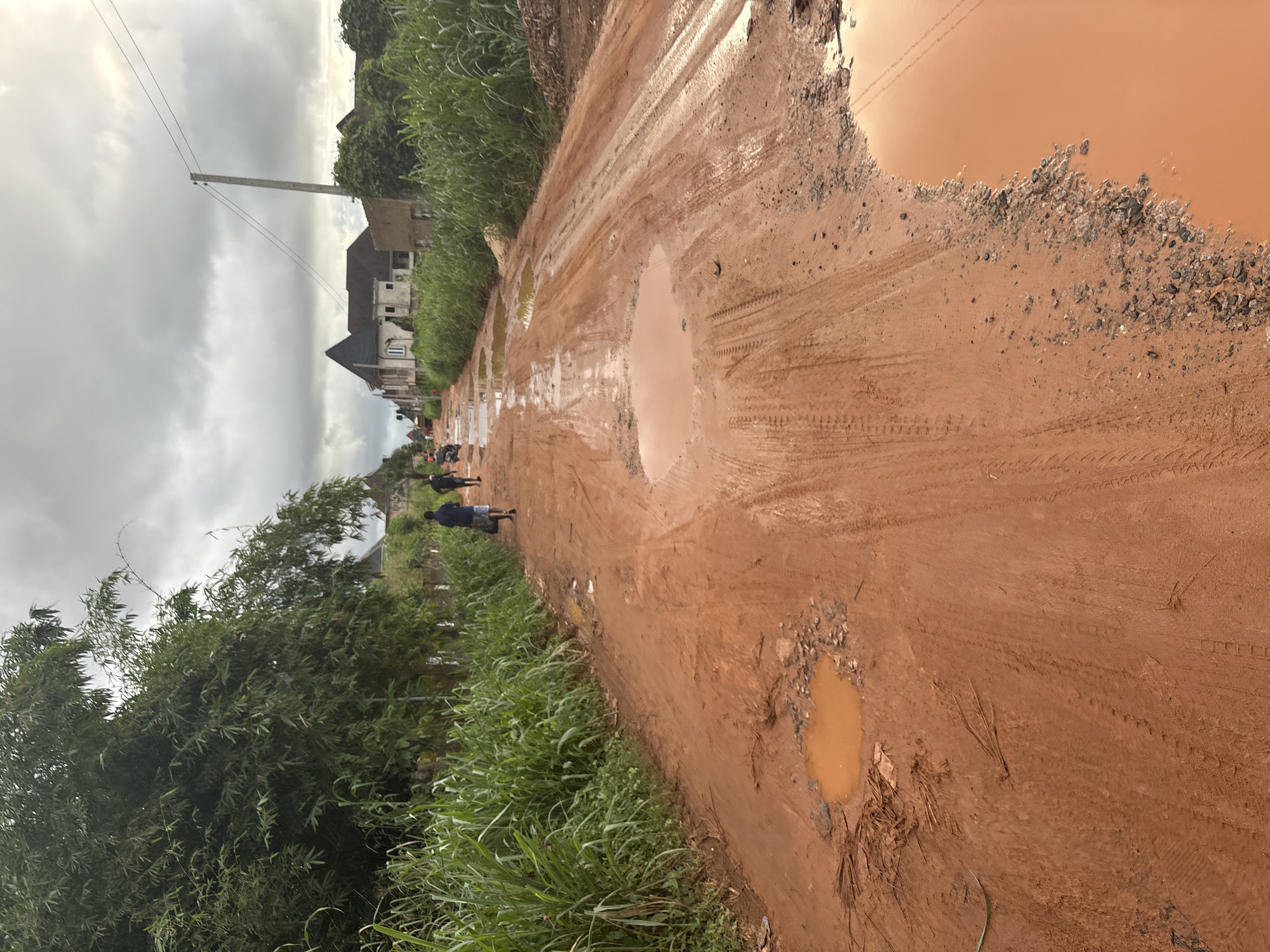 The image shows a muddy, unpaved road in Obosi, Nigeria. The road is covered in tire tracks and littered with puddles and debris. On the left side of the road, there is a dense growth of tall green grass and bamboo trees. Further down the road, several houses and buildings are visible, some appearing to be under construction. Two individuals are walking away from the camera on the road, and another person is on a motorbike in the distance. The sky is overcast, with grey clouds suggesting an impending storm. The scene depicts a rural or developing area with a challenging road infrastructure.