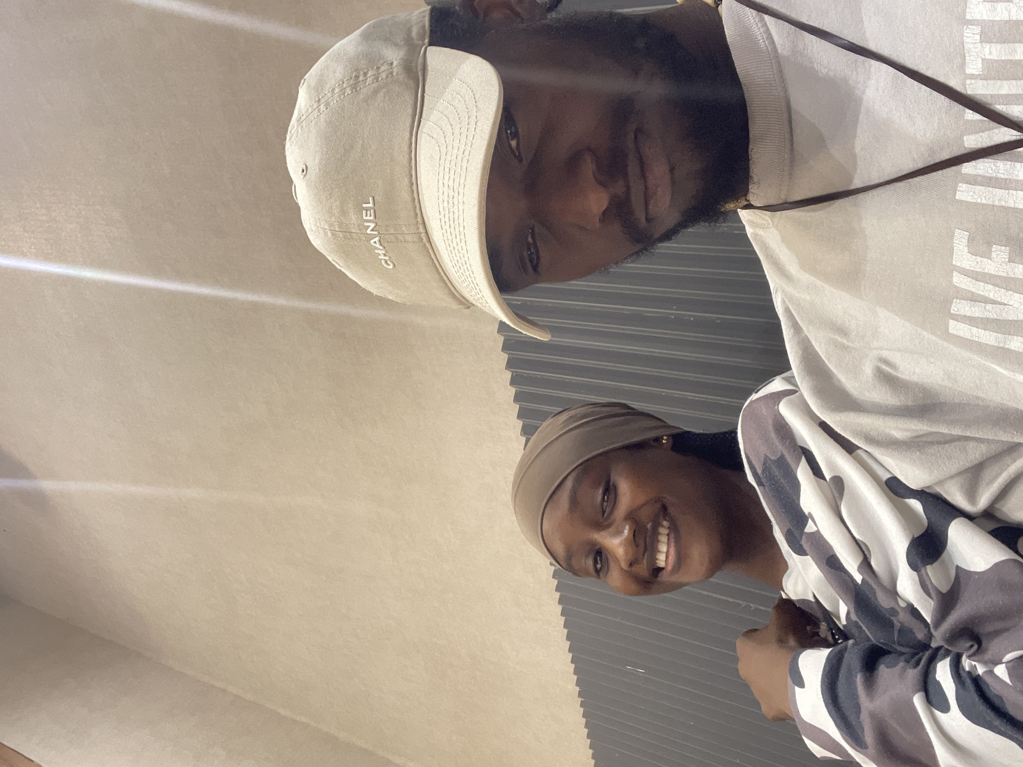 Two individuals, a woman and a man, are captured in a selfie. The woman, on the left, is wearing a brown headscarf and a camouflage-patterned sweatshirt. She is smiling and looking towards the camera. The man, on the right, is wearing a beige baseball cap with "CHANEL" embroidered on it, and a light-colored t-shirt. His expression is more neutral, and he is looking slightly away from the camera. The background appears to be an indoor setting with a textured, light-colored wall and a section of dark, ribbed paneling on the left. The lighting suggests an artificial light source, possibly overhead. The overall scene suggests a casual encounter or a shared moment between the two individuals. The image does not contain enough visual information to determine the specific location in Guzape, Nigeria.