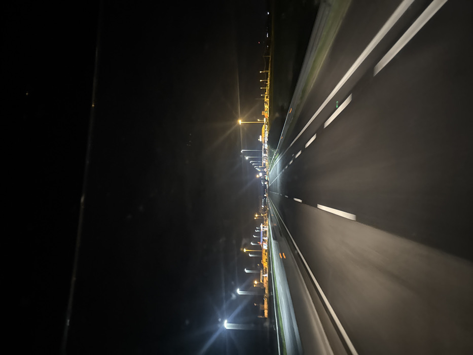 This image depicts a nighttime scene from the perspective of a vehicle traveling on a multi-lane highway near Foggia, Italy. The asphalt roadway, illuminated by bright white streetlights, features distinct solid and dashed white lane markings, with at least two lanes visible in the direction of travel. A concrete or metal guardrail borders the left side of the highway, suggesting a median, and another runs along the right shoulder. In the mid-ground, extending to the left and right sides of the highway, are concentrations of yellow and orange lights, indicating the presence of commercial or industrial structures, possibly a service area or complex. The sky above is dark, characteristic of nighttime conditions. A faint, thin horizontal line, possibly an overhead wire or part of the vehicle's interior, traverses the upper portion of the frame. No specific people or other vehicles are distinctly identifiable within the frame, but the scene implies active travel.
